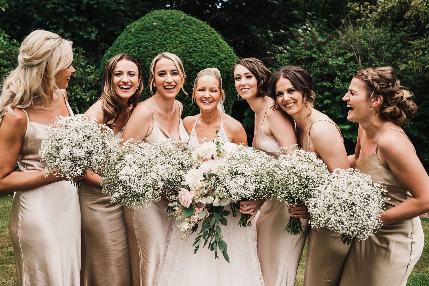 Group of bridesmaids in champagne dresses holding baby's breath bouquets pose together outdoors.