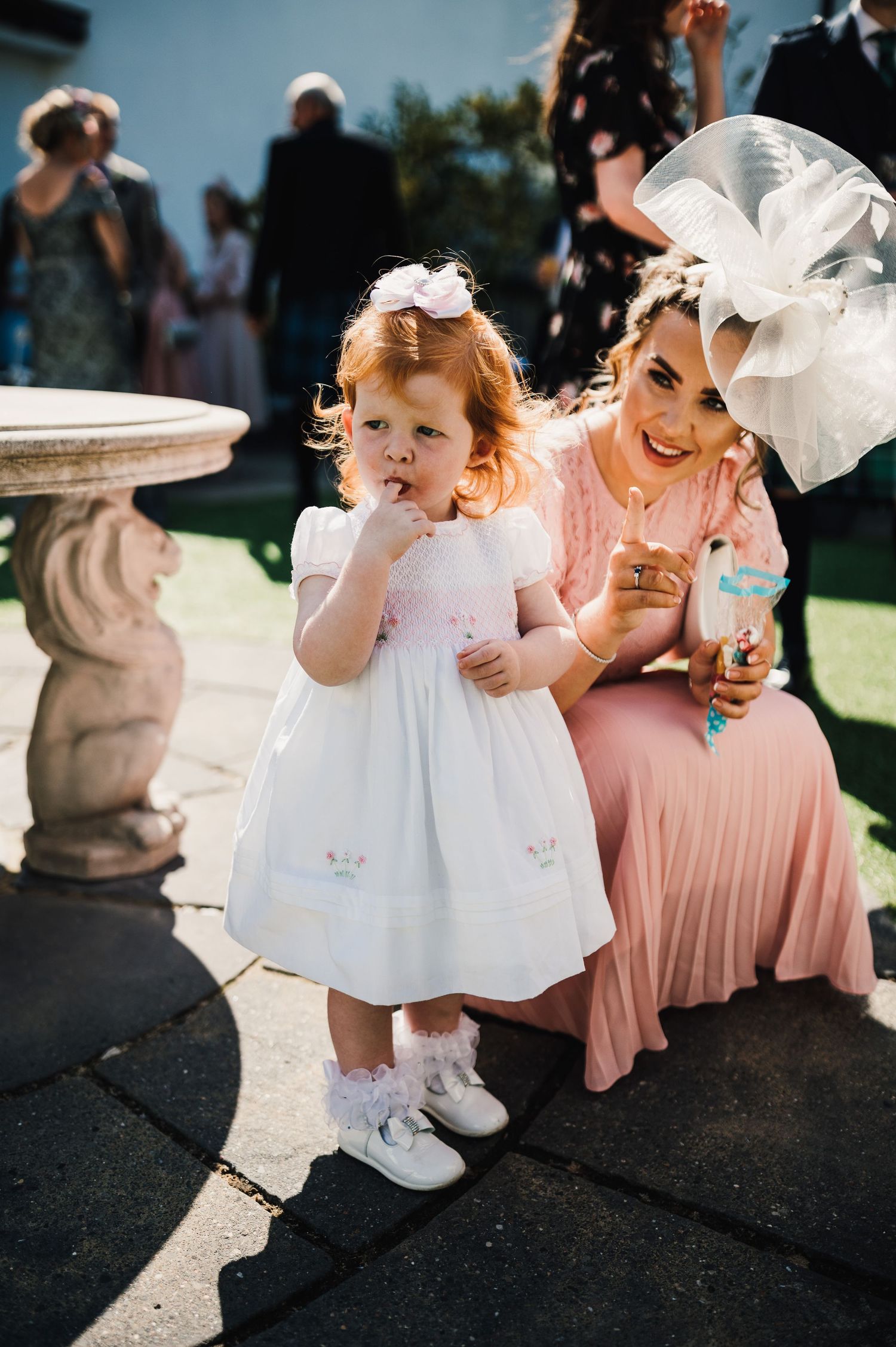 Young guest in white dress enjoys a snack at an outdoor wedding reception next to woman in pink dress.
