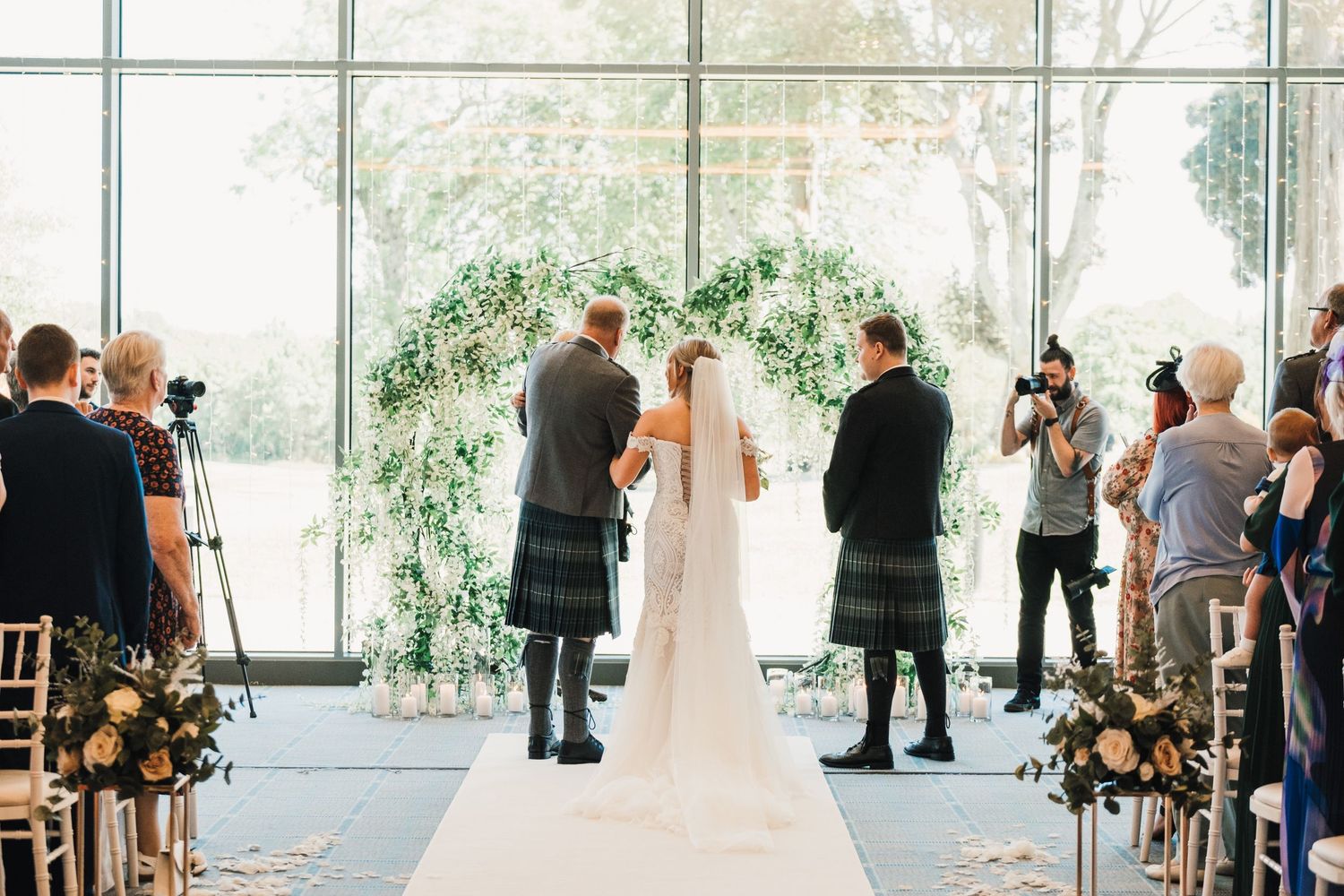 Wedding ceremony in bright modern venue with Scottish kilts and floral decorations.