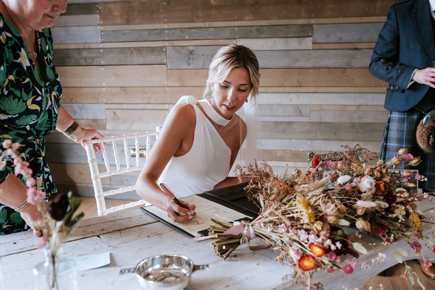 Couple laughing while completing wedding paperwork at wooden table with autumn flowers.