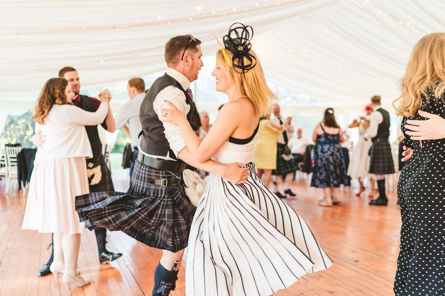 Dancers moving across a polished dance floor under a white tent during a festive gathering.