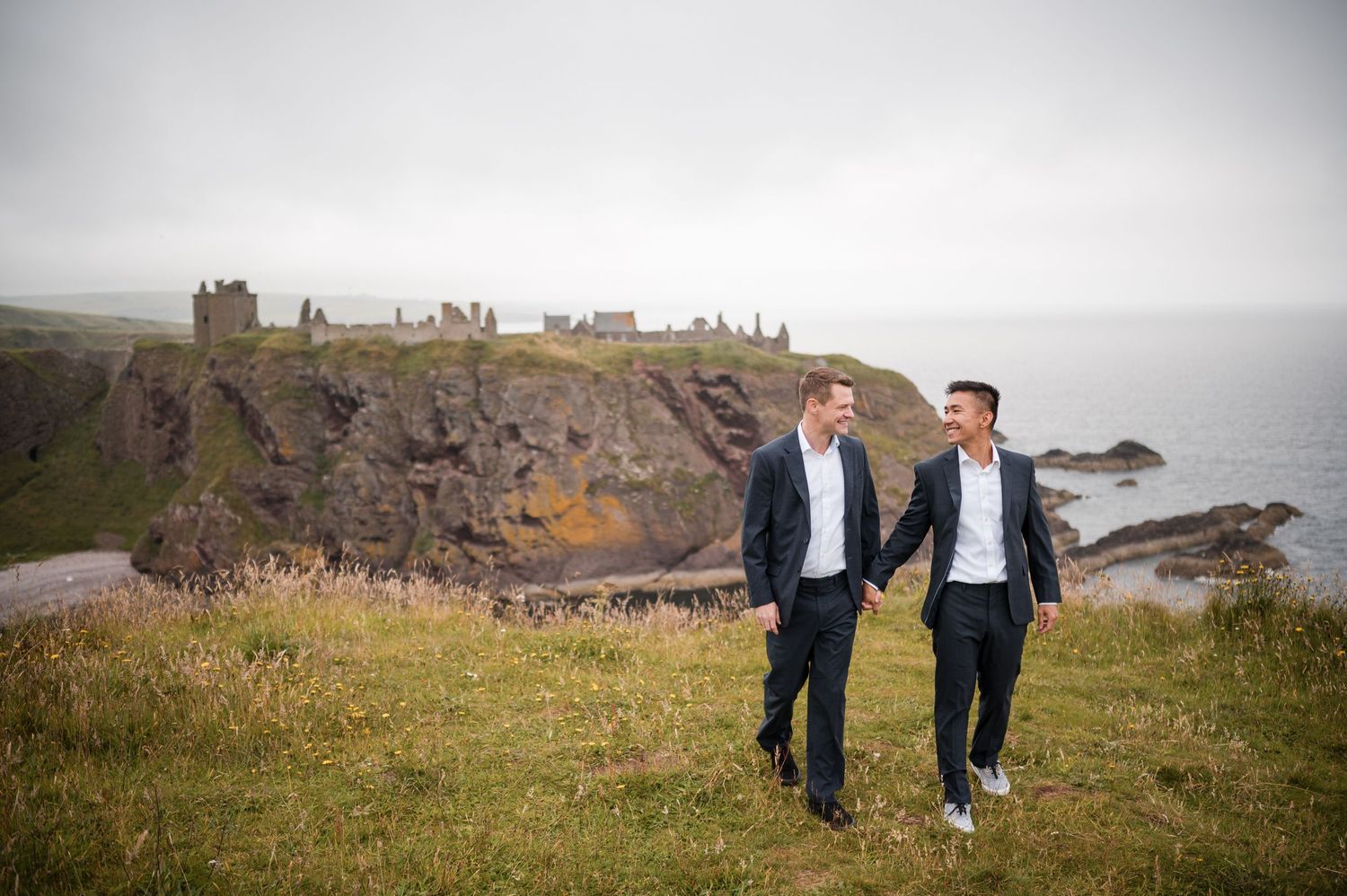 Same Sex Wedding PhotographA couple walks hand in hand along a clifftop path with dramatic coastal views and historic ruins in the distance.