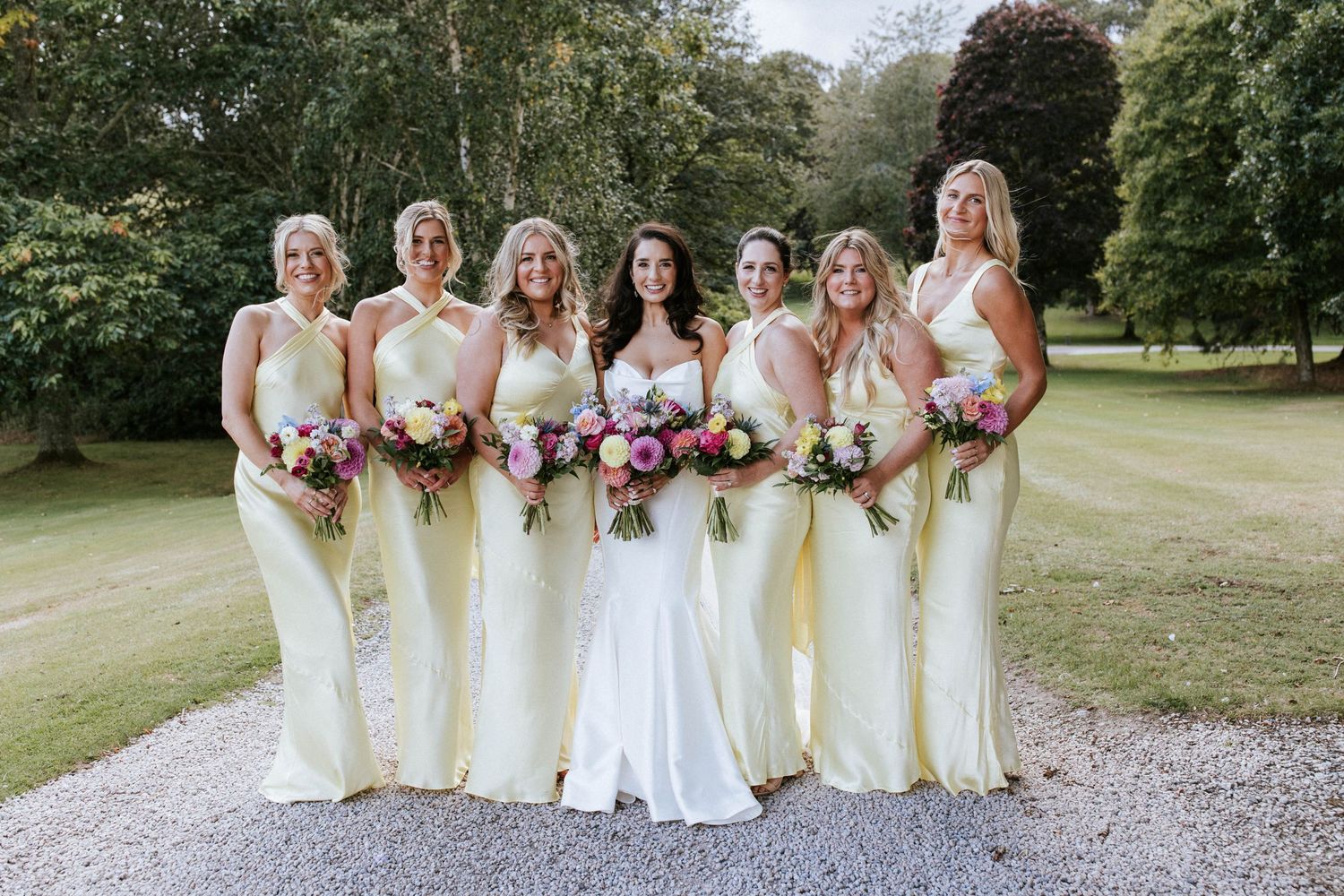 Bridal party in elegant yellow gowns poses with bouquets on a gravel path surrounded by greenery.