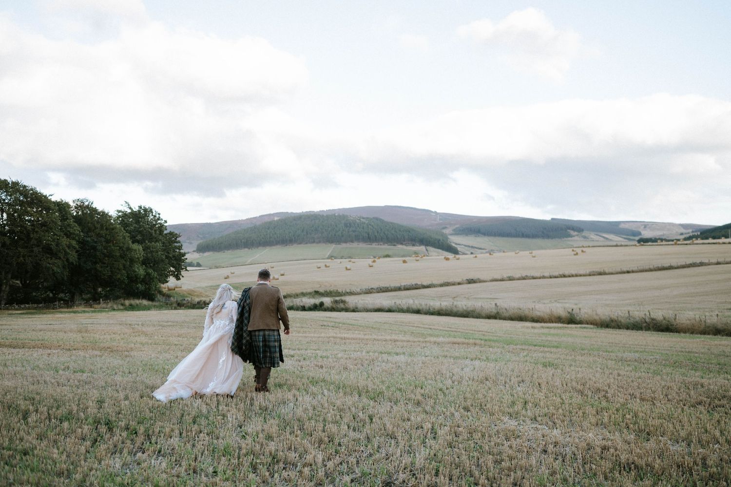 Couple walking through vast countryside field under cloudy sky.