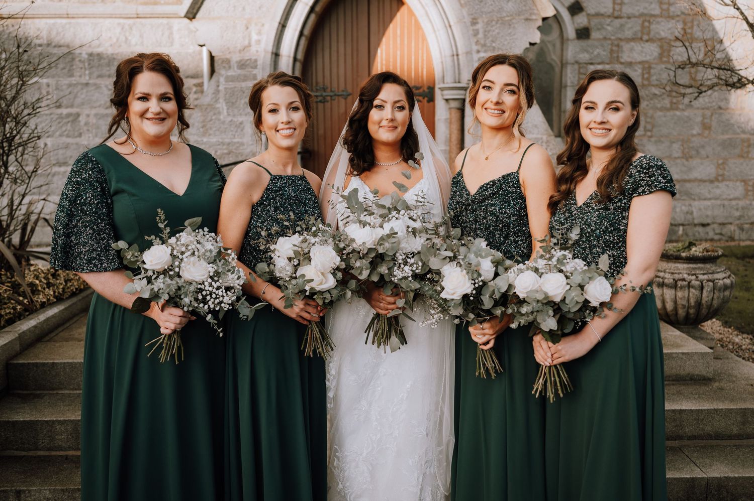 Bridal party in dark green sequined and solid dresses hold winter bouquets outside church.