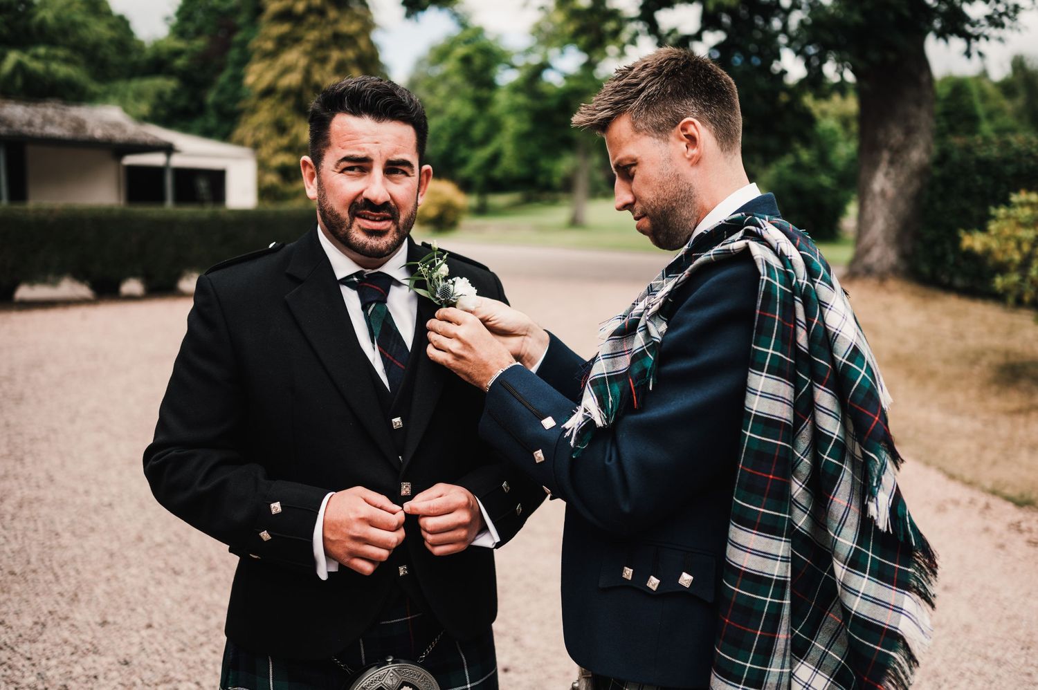 Two men in Scottish formal wear share a laugh while standing on gravel path.