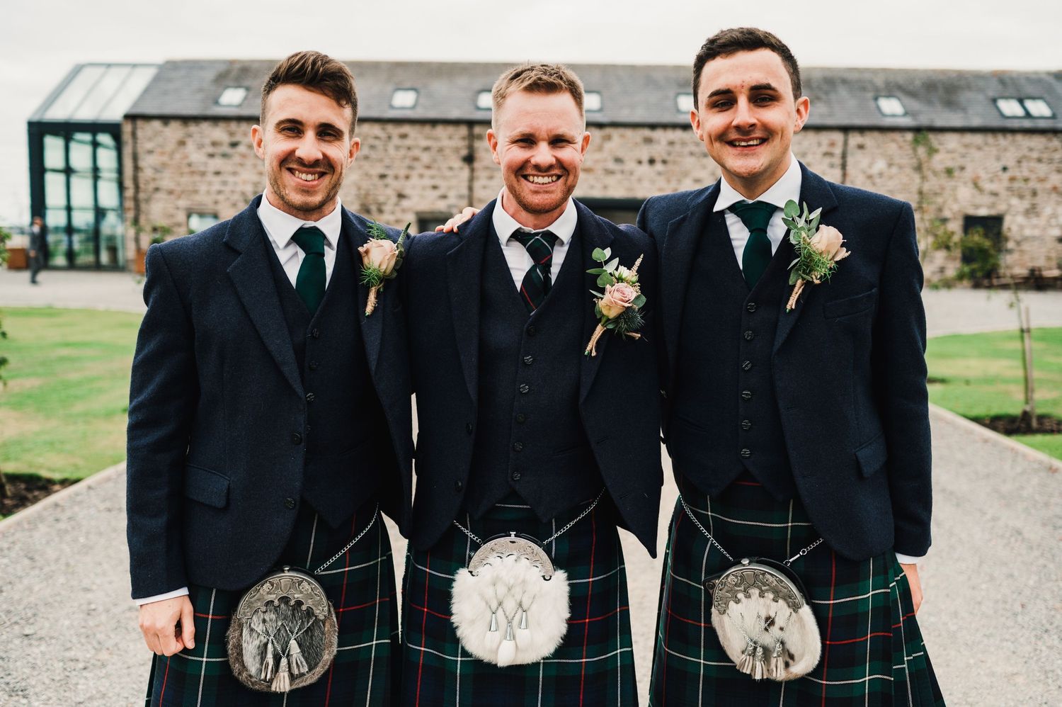 Three groomsmen in matching kilts and jackets smile at a Scottish wedding venue.