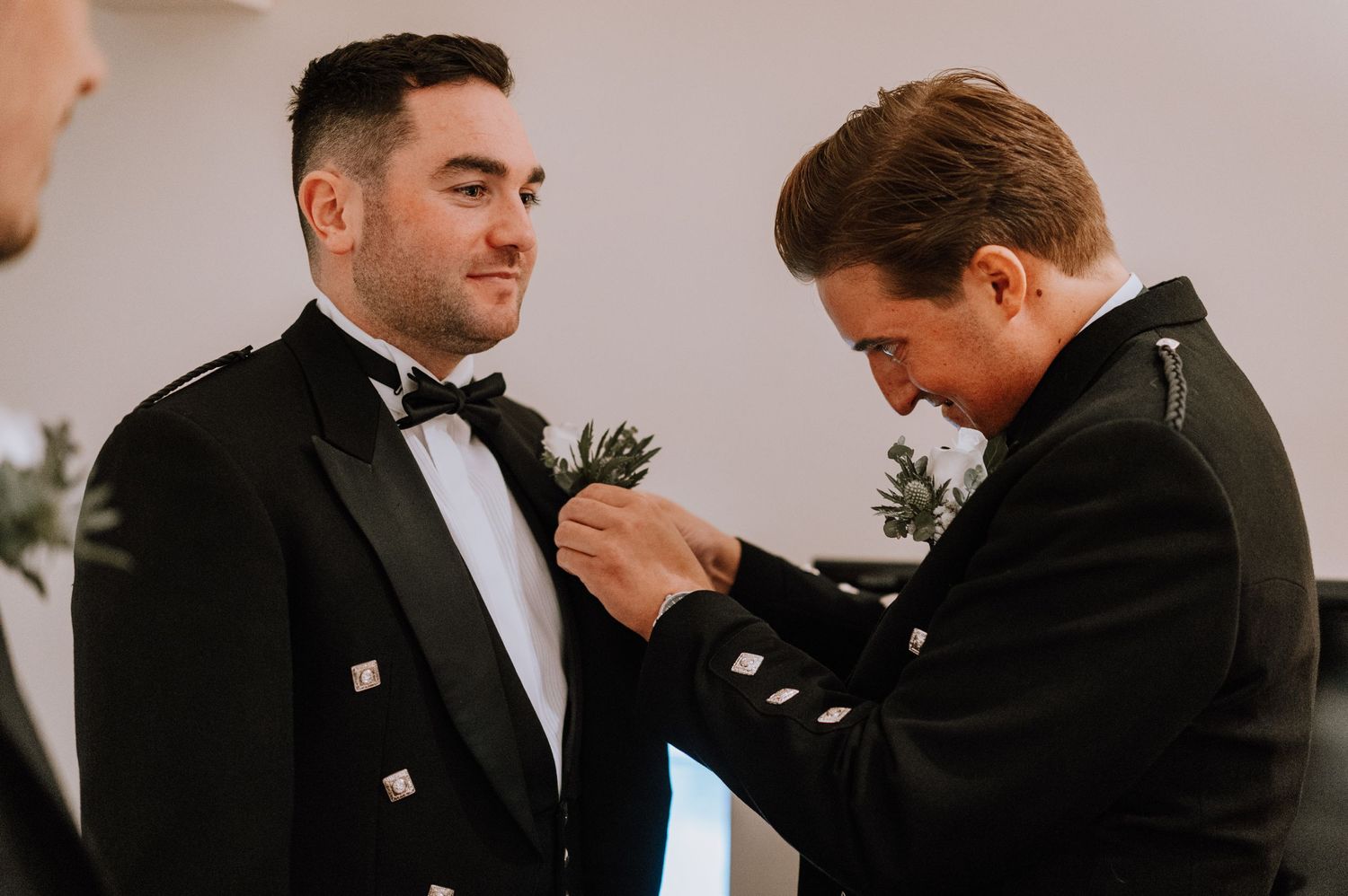 Groomsmen in traditional Scottish formal wear helping with boutonniere pinning.
