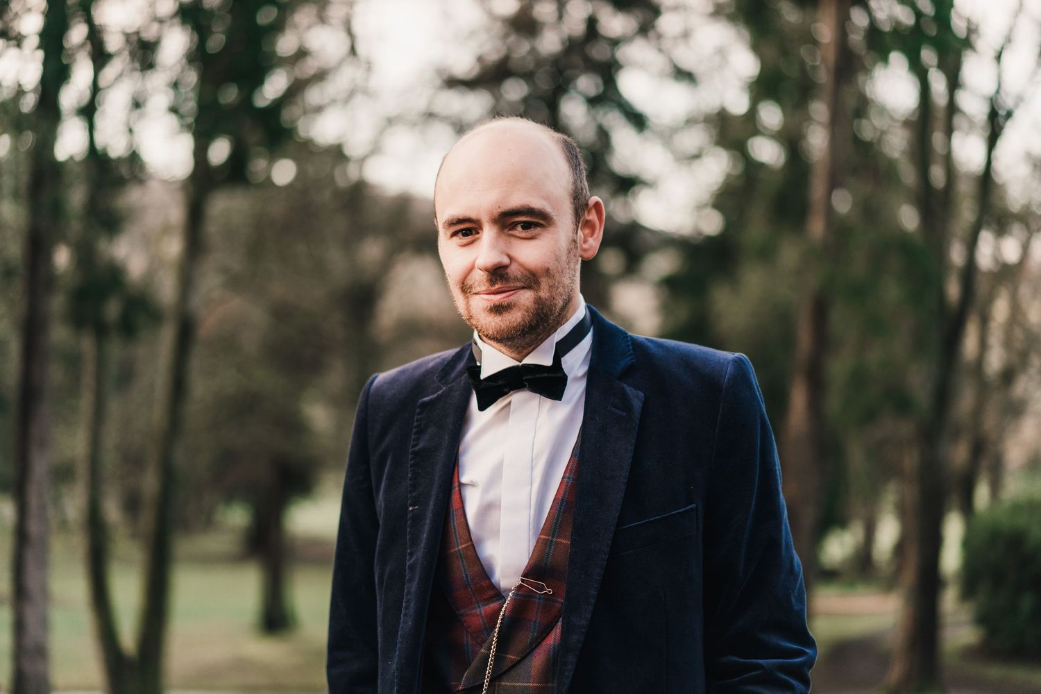 A groom wearing a navy suit with burgundy vest and bowtie smiles while standing outdoors among trees.