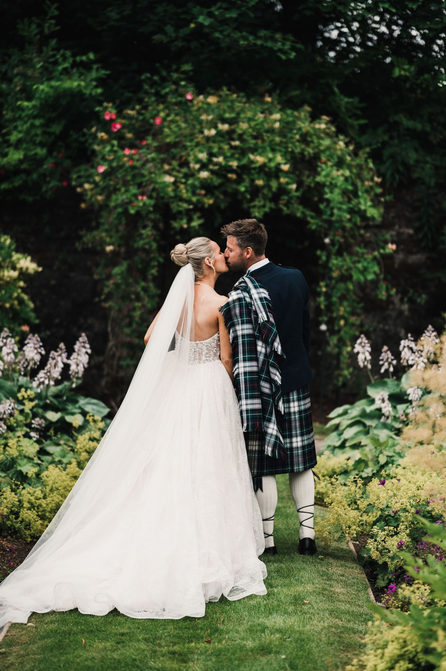 Bride and groom share an intimate moment in a garden, with the groom wearing traditional Scottish attire.