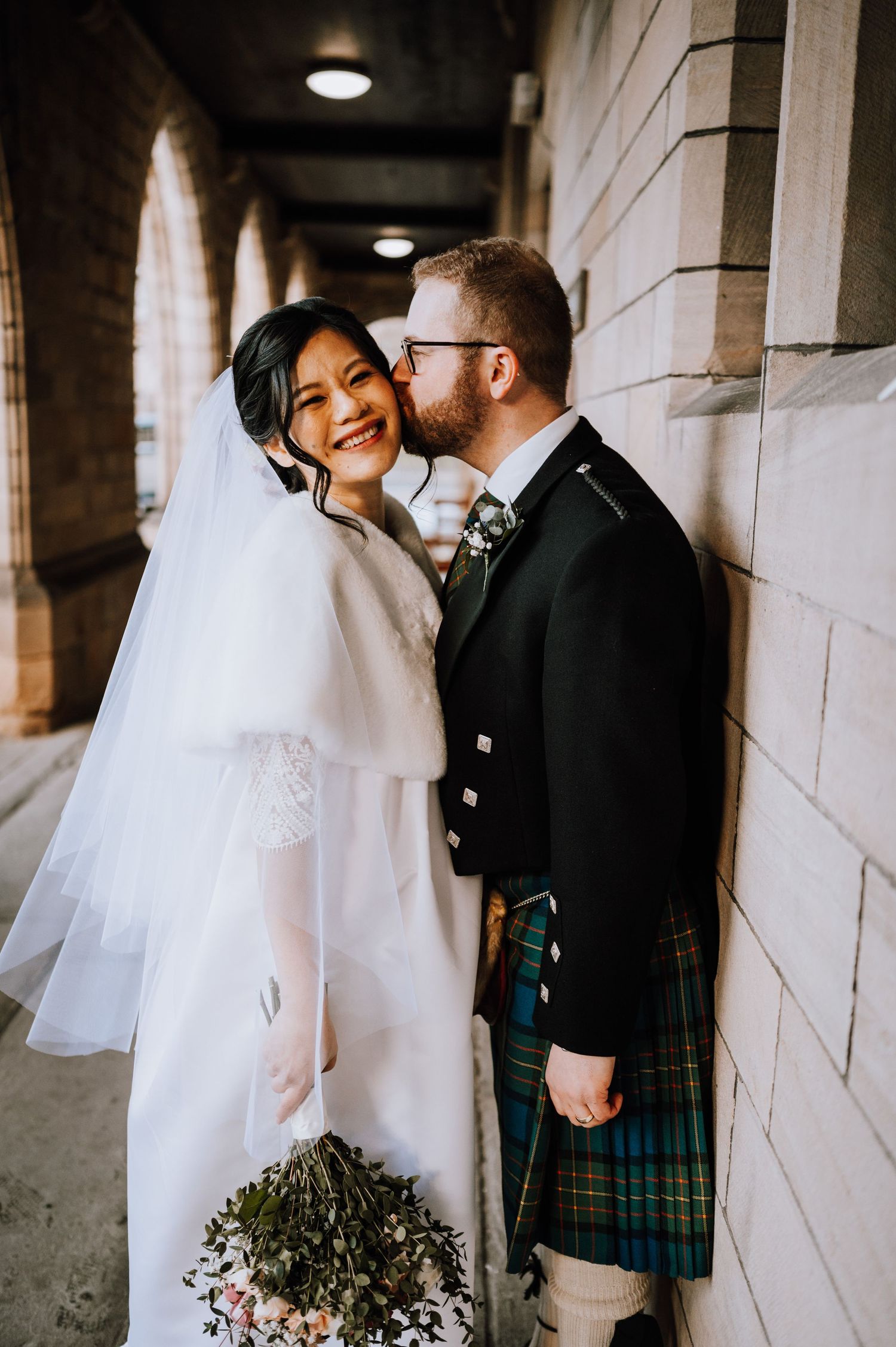 Wedding couple stands beneath Gothic church archway in white dress and Scottish highland wear.