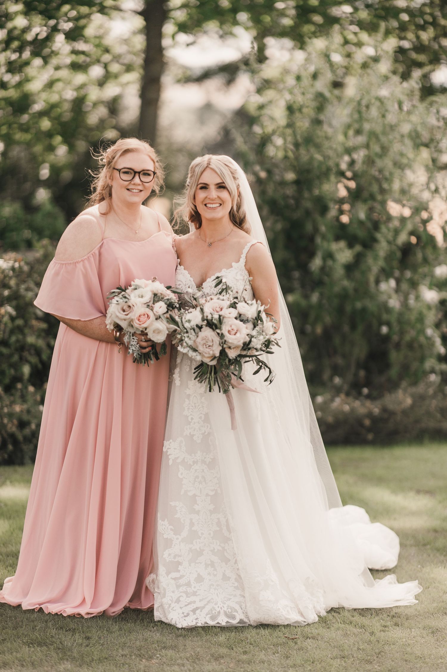 Wedding party pose outdoors with pink bridesmaid dress and white lace bridal gown holding neutral floral bouquets.