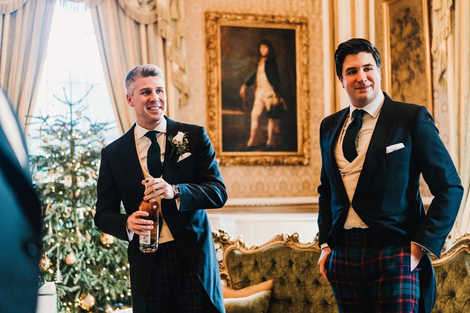 Two groomsmen in formal suits enjoying drinks in an ornate room with traditional decor and paintings.