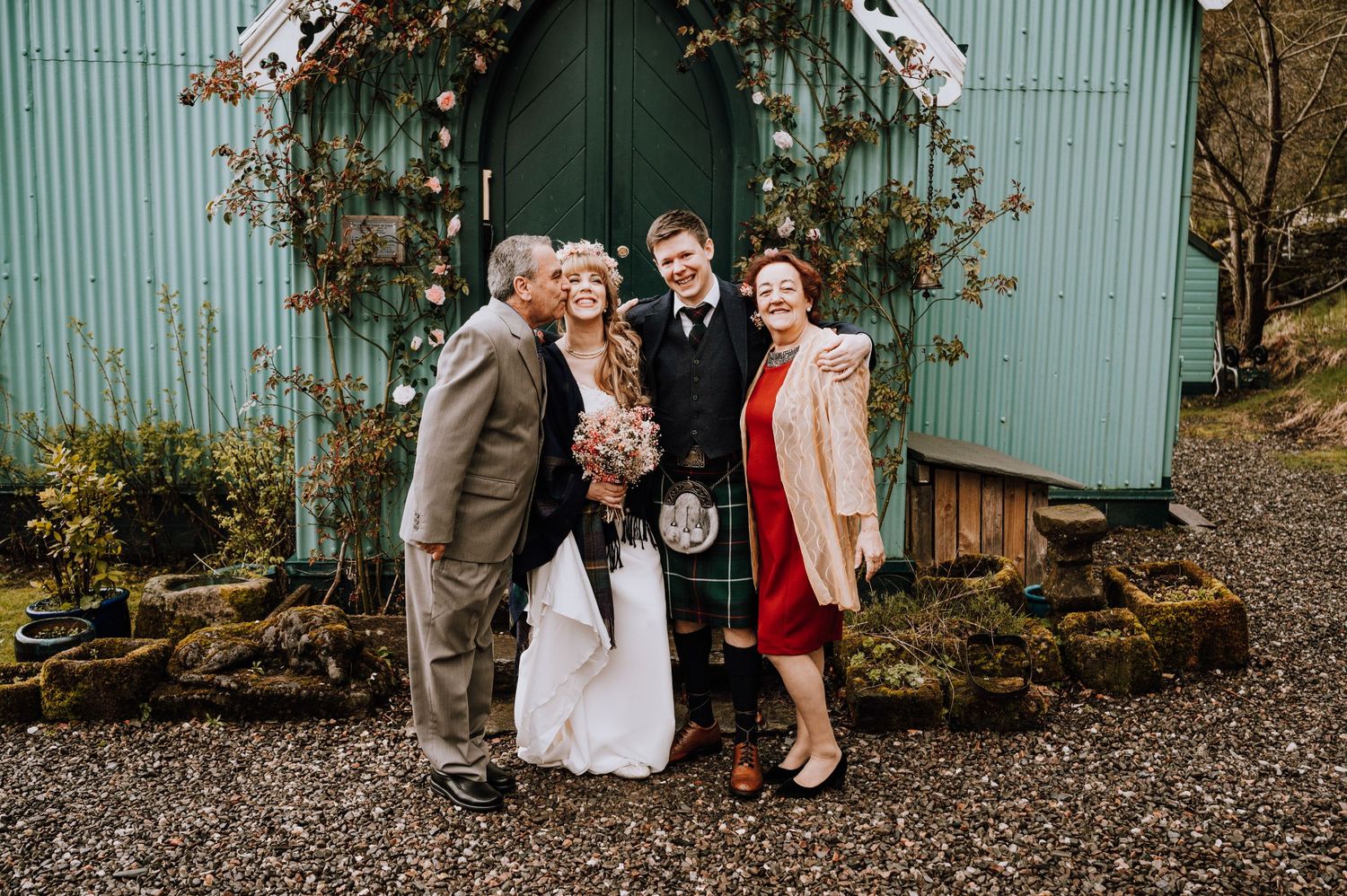 Wedding photo of group standing in front of teal church with floral archway.