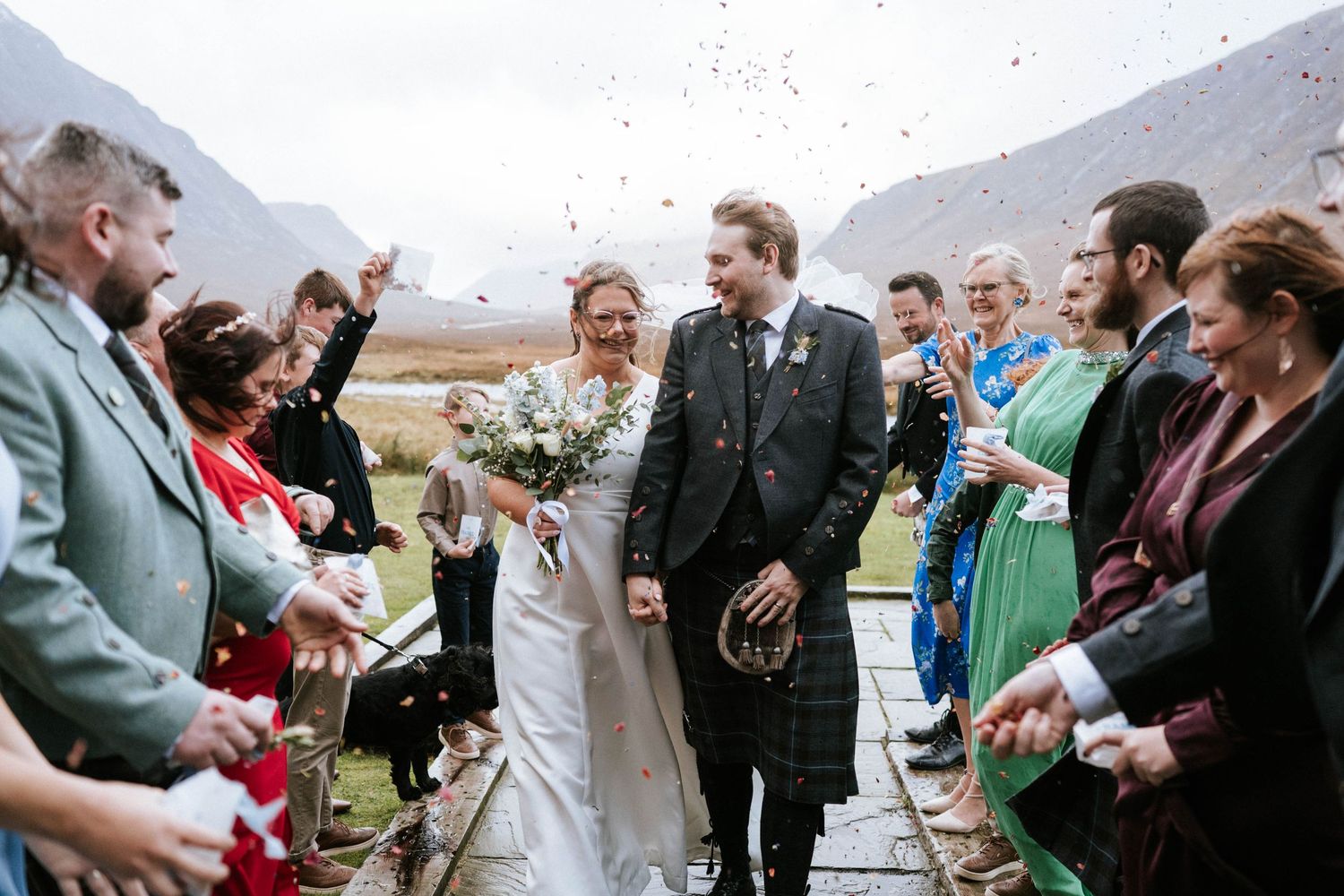 Wedding guests celebrate with confetti and cheers outside in a scenic mountain landscape.