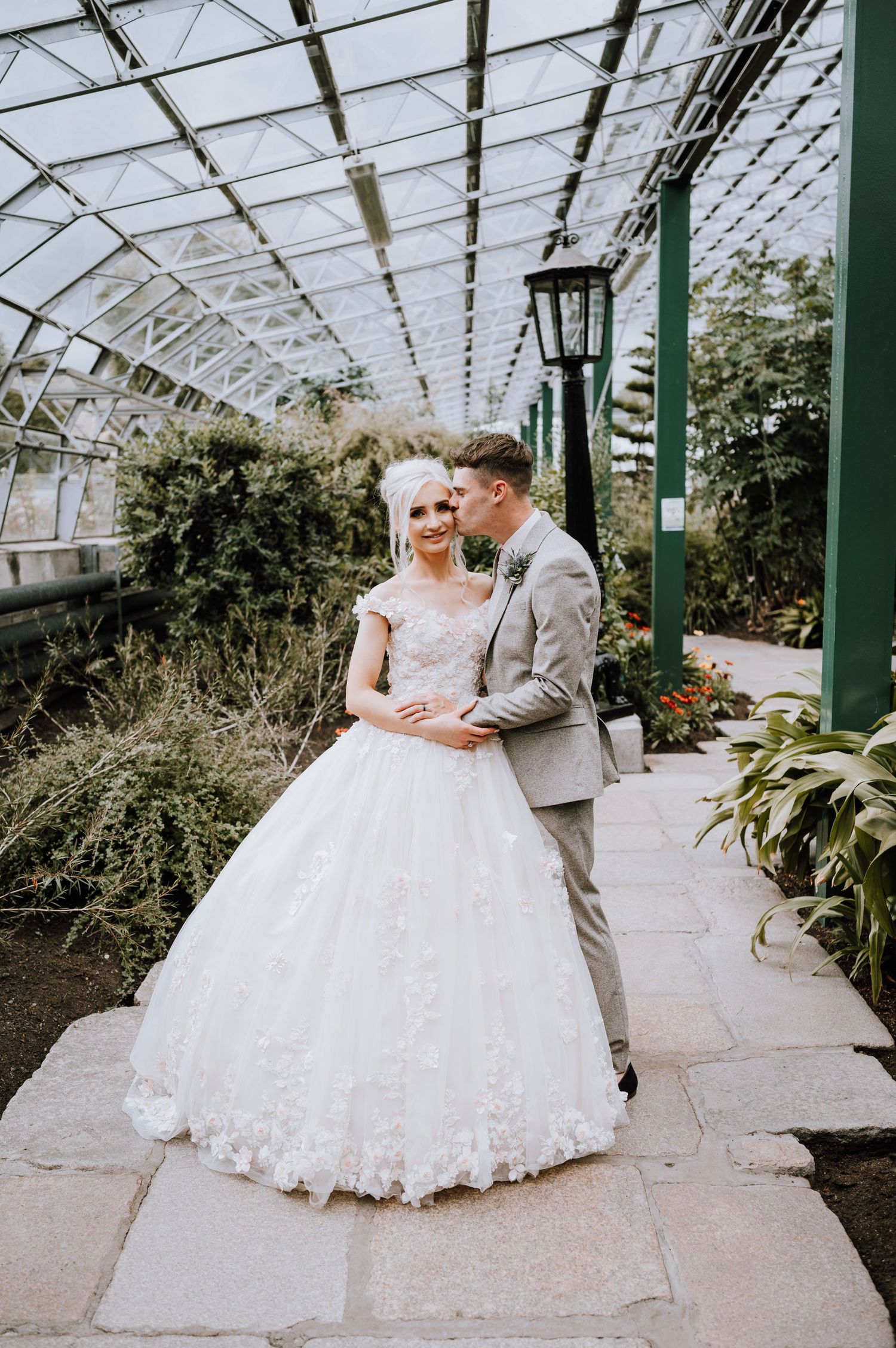 A couple poses for wedding photos in an elegant greenhouse with hanging lanterns.