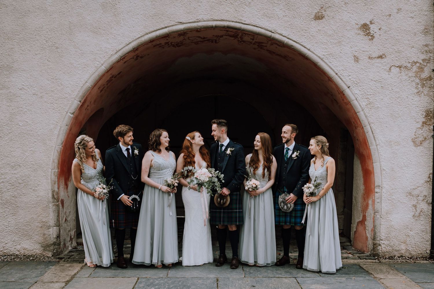 A bridal party stands together under a rustic stone archway for a formal wedding photo.