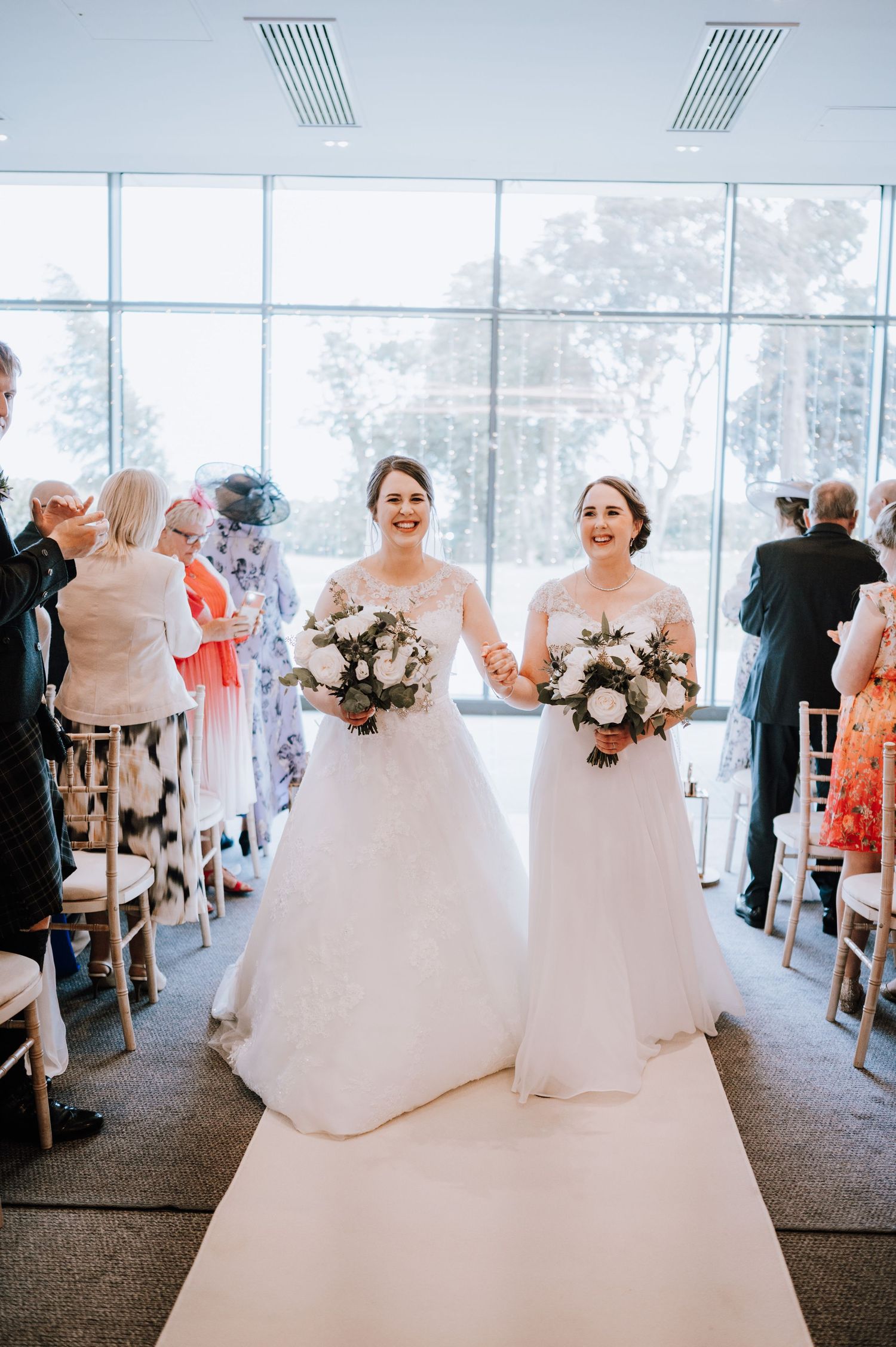 Two people in wedding attire hold bouquets while walking down a white aisle runner surrounded by guests.