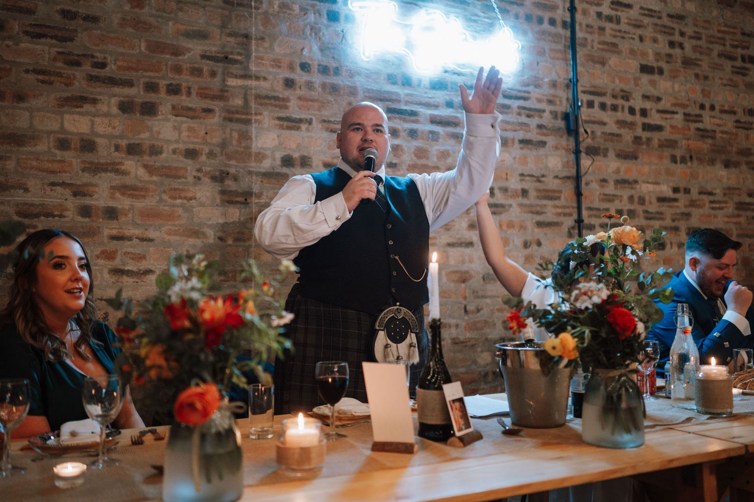A figure stands speaking and gesturing in formal attire at a reception table decorated with floral arrangements and candles.