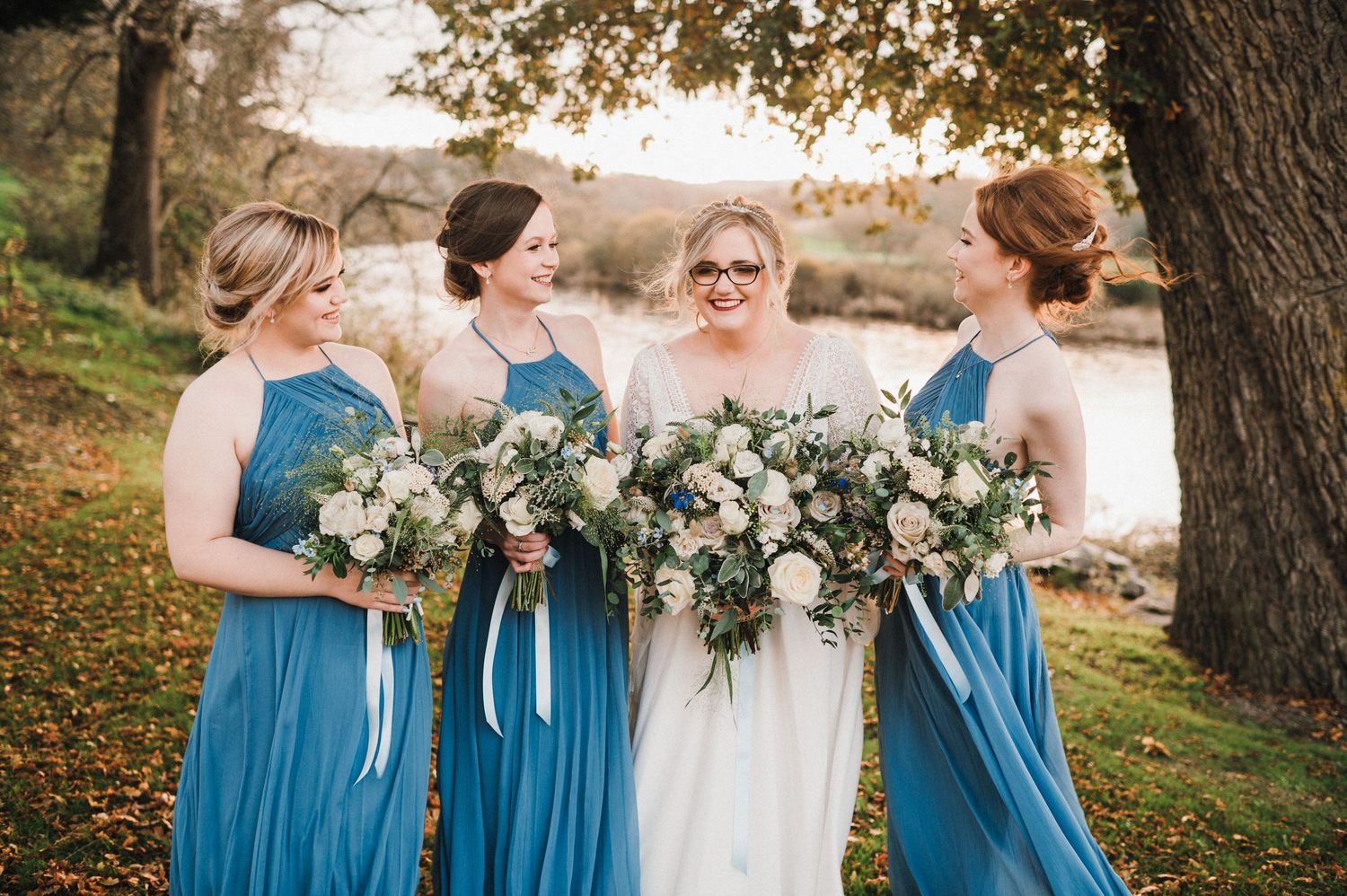 Bridesmaids in flowing blue dresses hold white floral bouquets while gathered outdoors.
