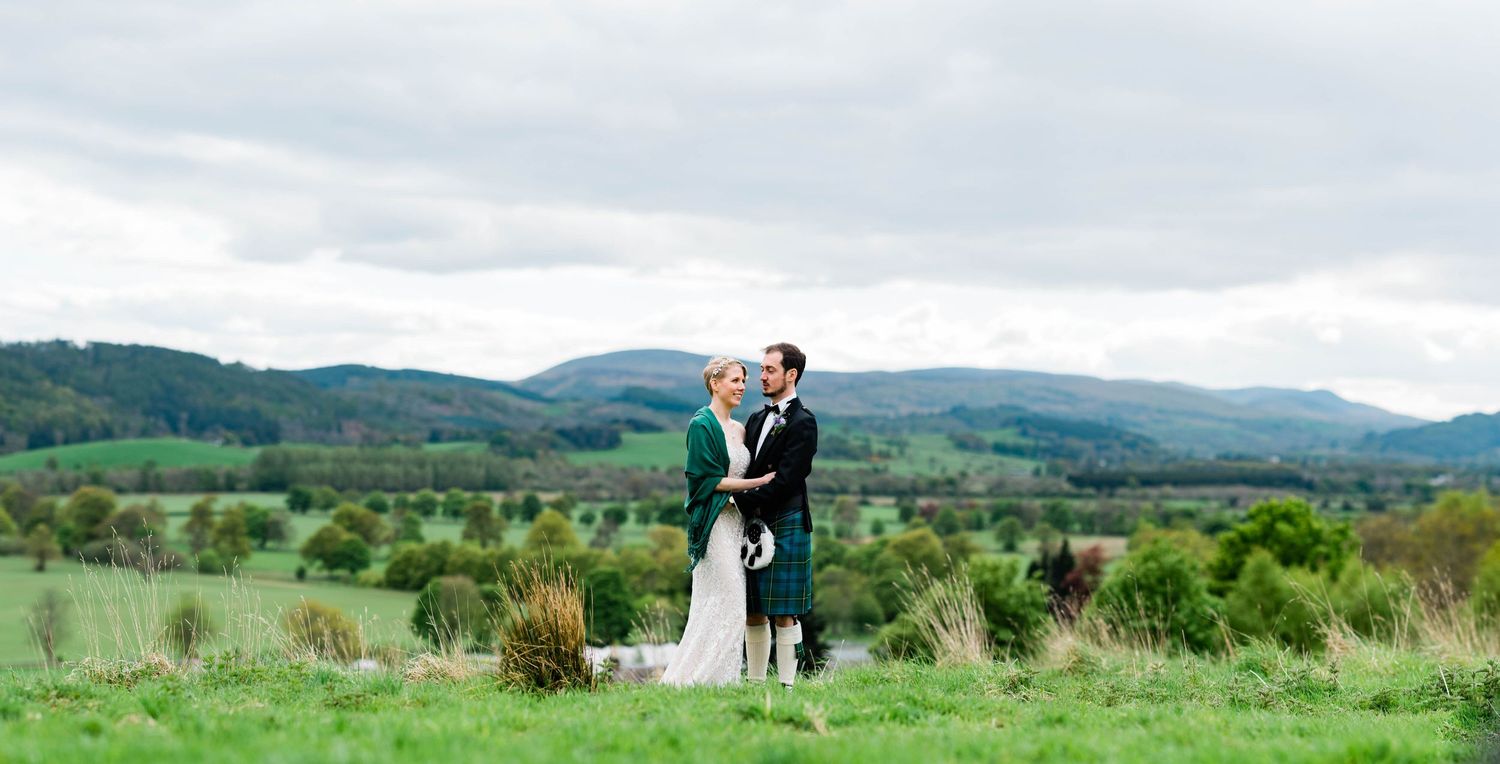 A wedding couple embraces on a hilltop with rolling Scottish highlands in the background.