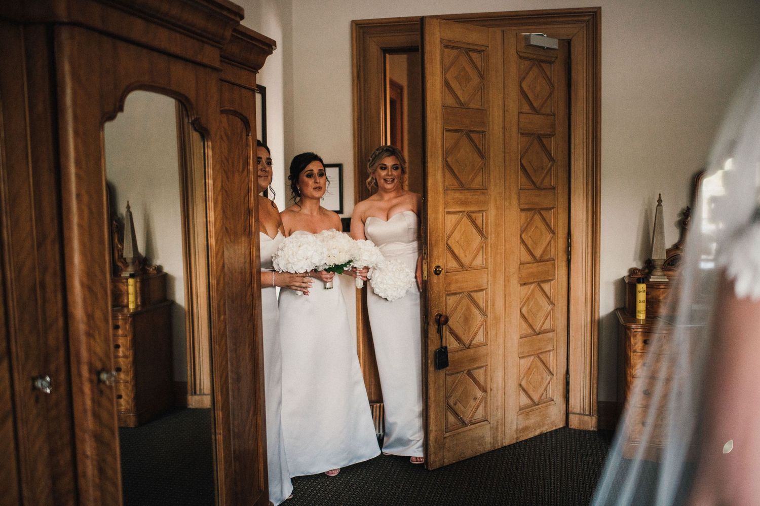 Two people in white dresses share a moment in an elegant room with wooden doors.