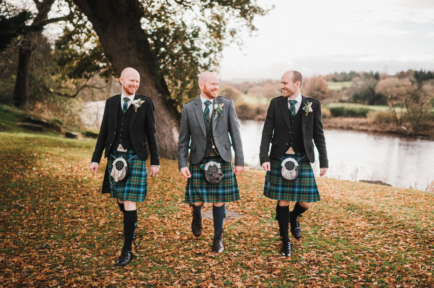 Three men in traditional Scottish kilts walking together on fallen autumn leaves by a lake.