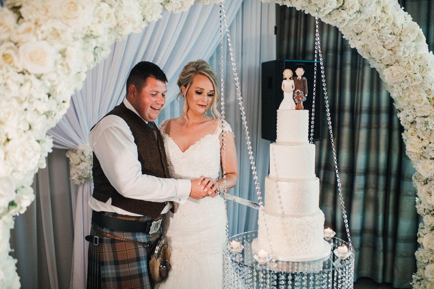 Couple admiring their elegant white wedding cake under floral arch.