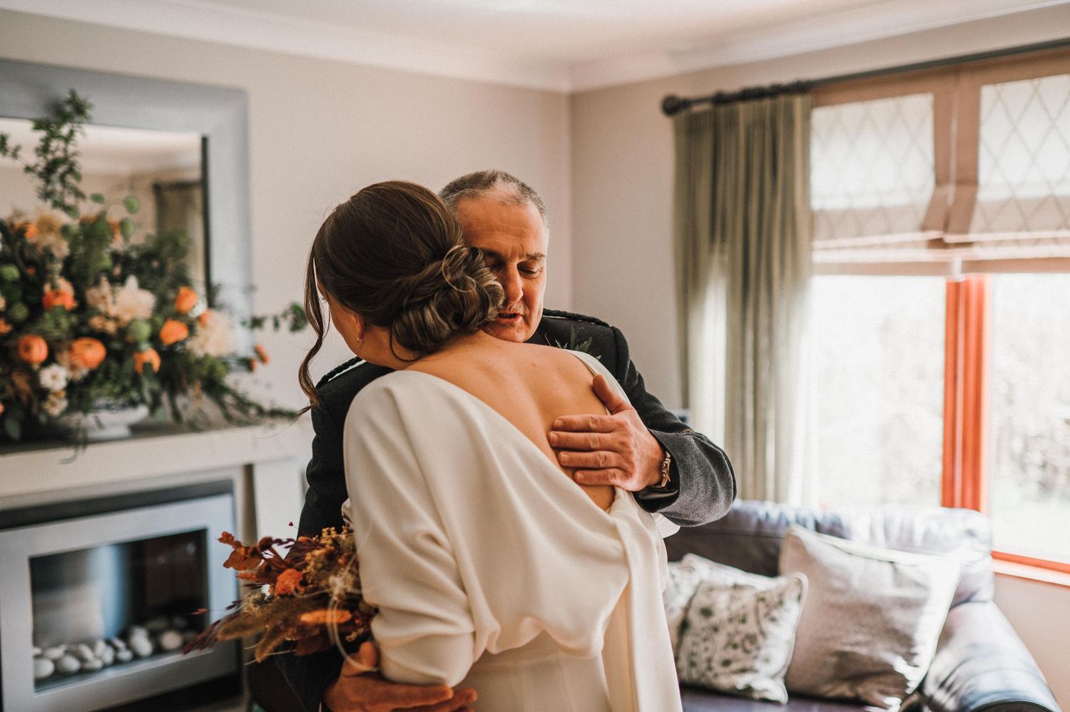 Two people share an emotional embrace in a well-decorated living room with peach floral arrangements.