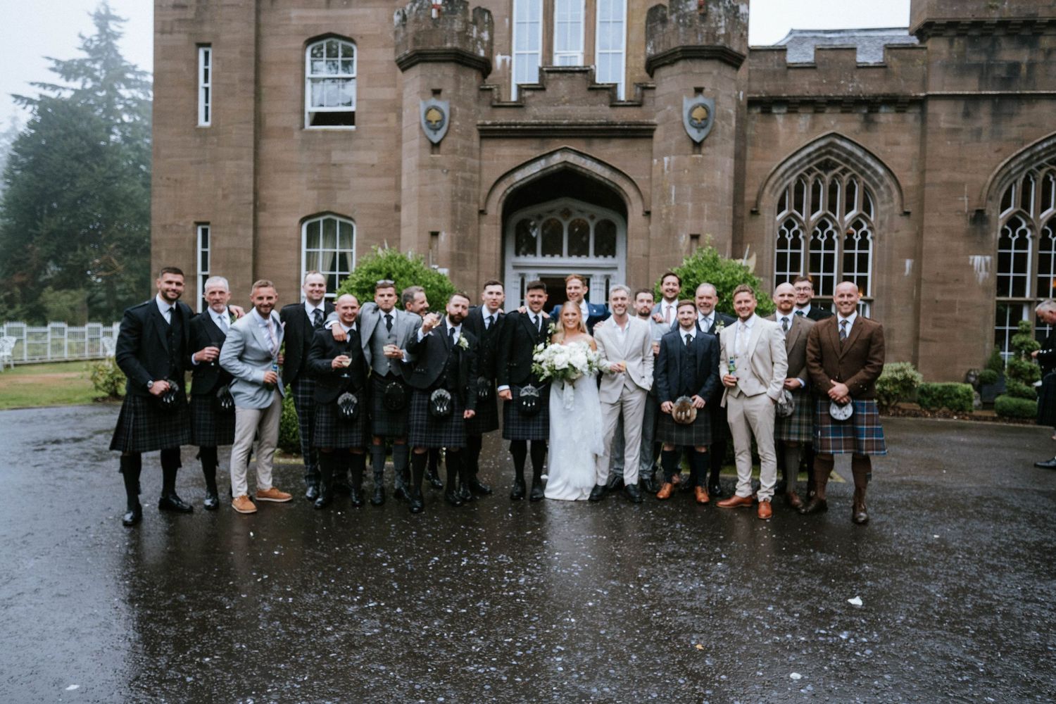 Wedding party group photo in front of a stone Gothic church building on a rainy day.