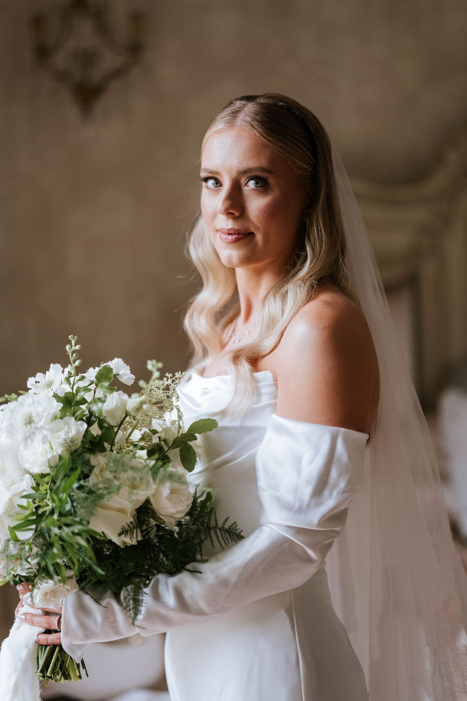 Elegant bride in an off-shoulder white wedding gown holds a lush bouquet of white flowers and greenery.