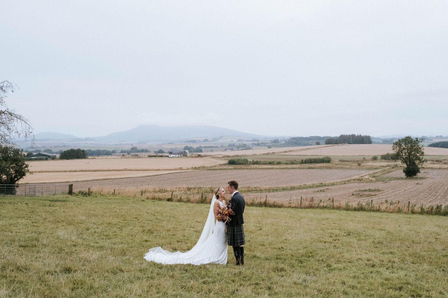 Newlyweds walking together in scenic countryside field on wedding day.