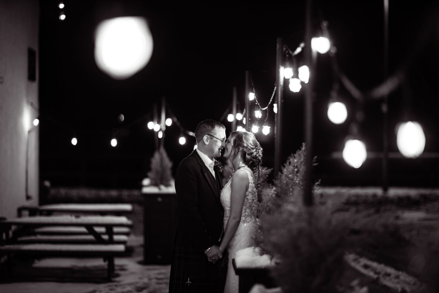 A romantic outdoor evening setting with string lights illuminating a dark patio area and picnic tables.