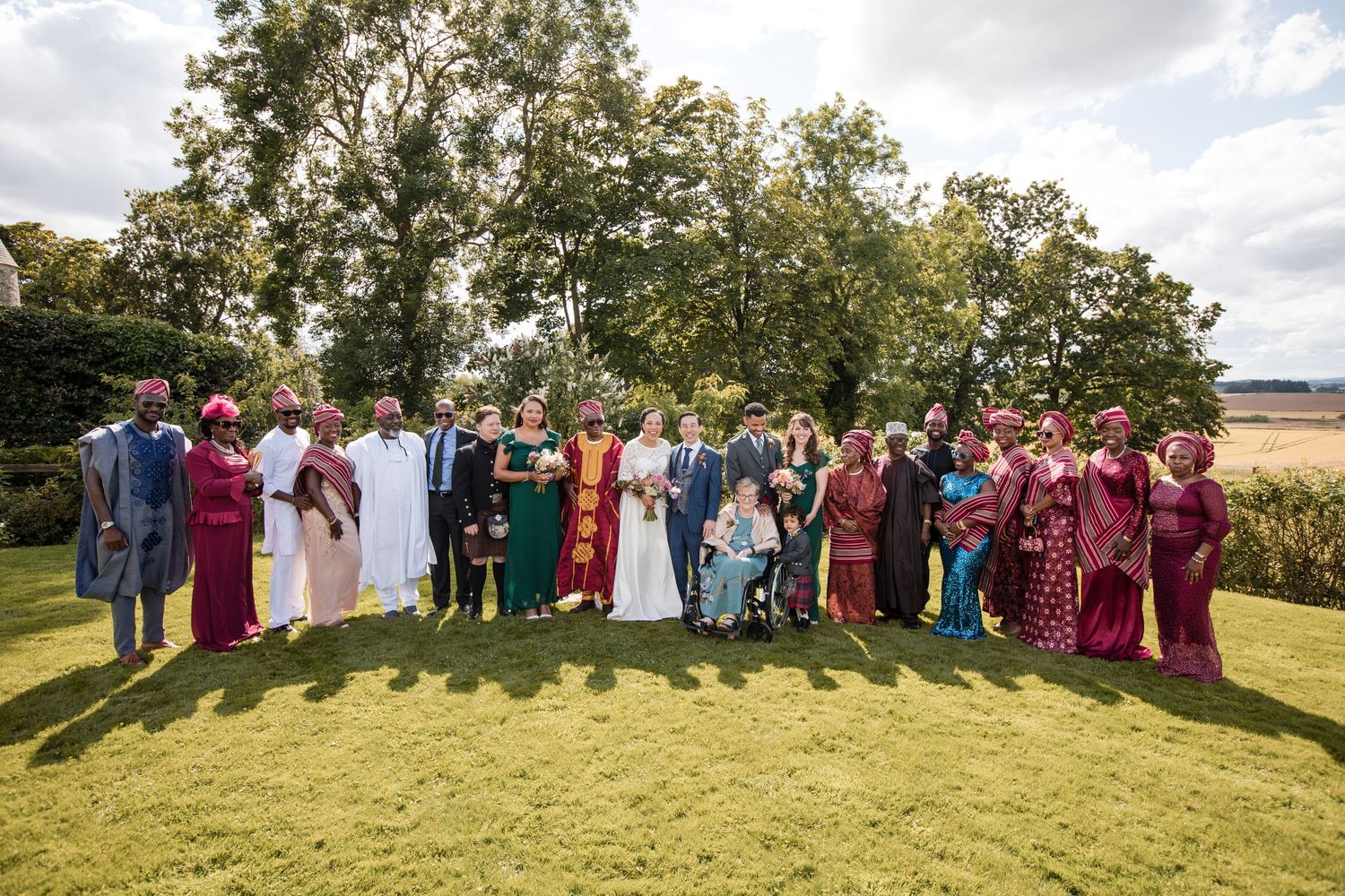 Large group wedding photo with guests arranged on grassy hillside under cloudy skies.