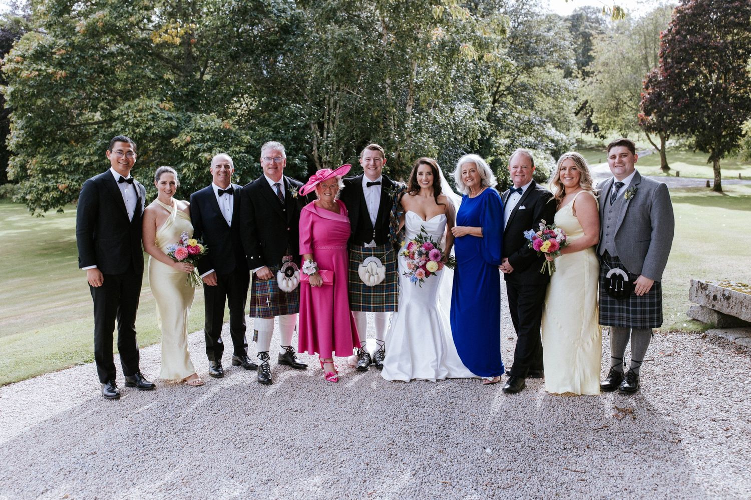 Extended wedding party in formal wear and Scottish kilts gathered on garden path for group photo.