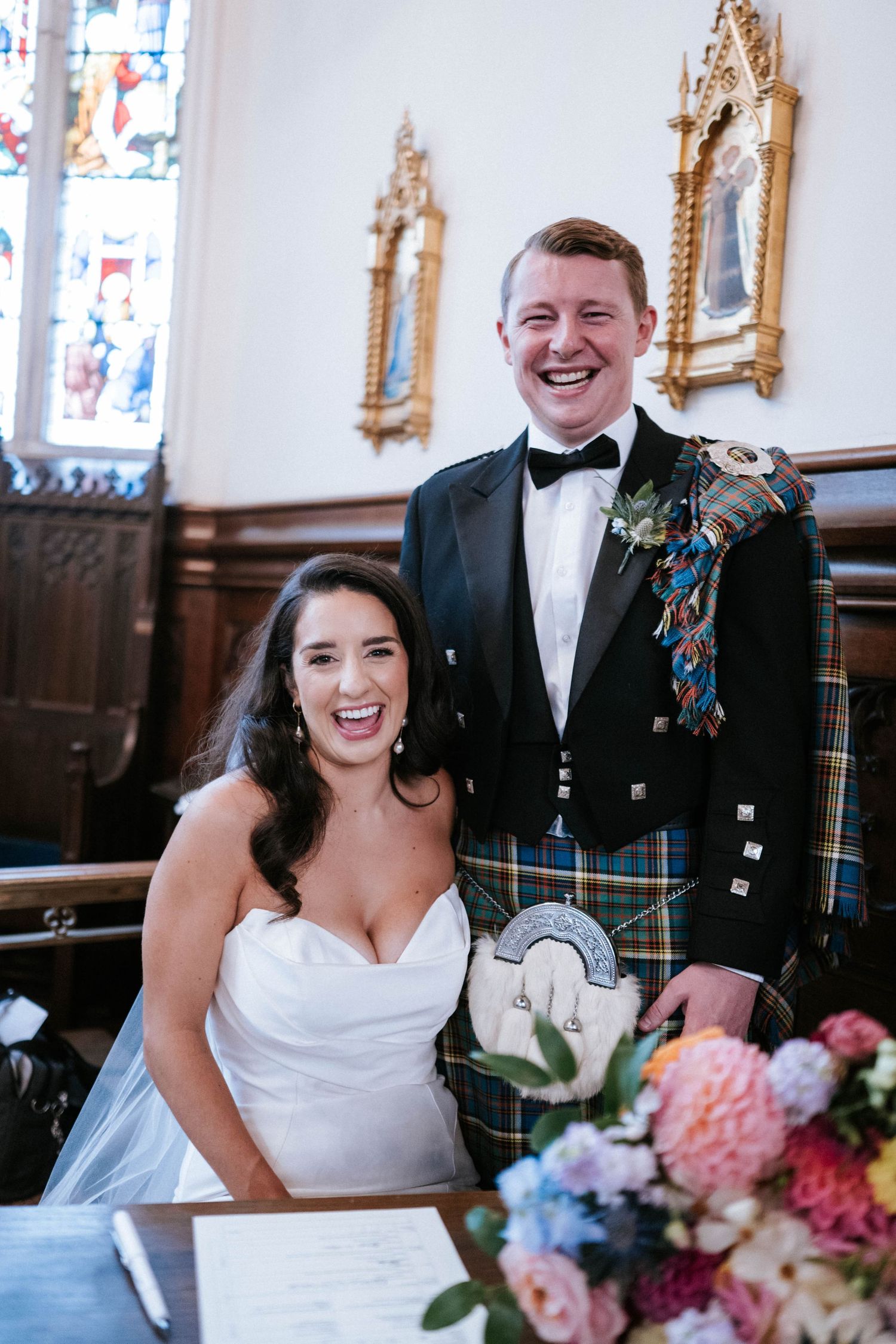 Joyful wedding couple posing at signing table with traditional Scottish kilt and elegant white bridal gown.