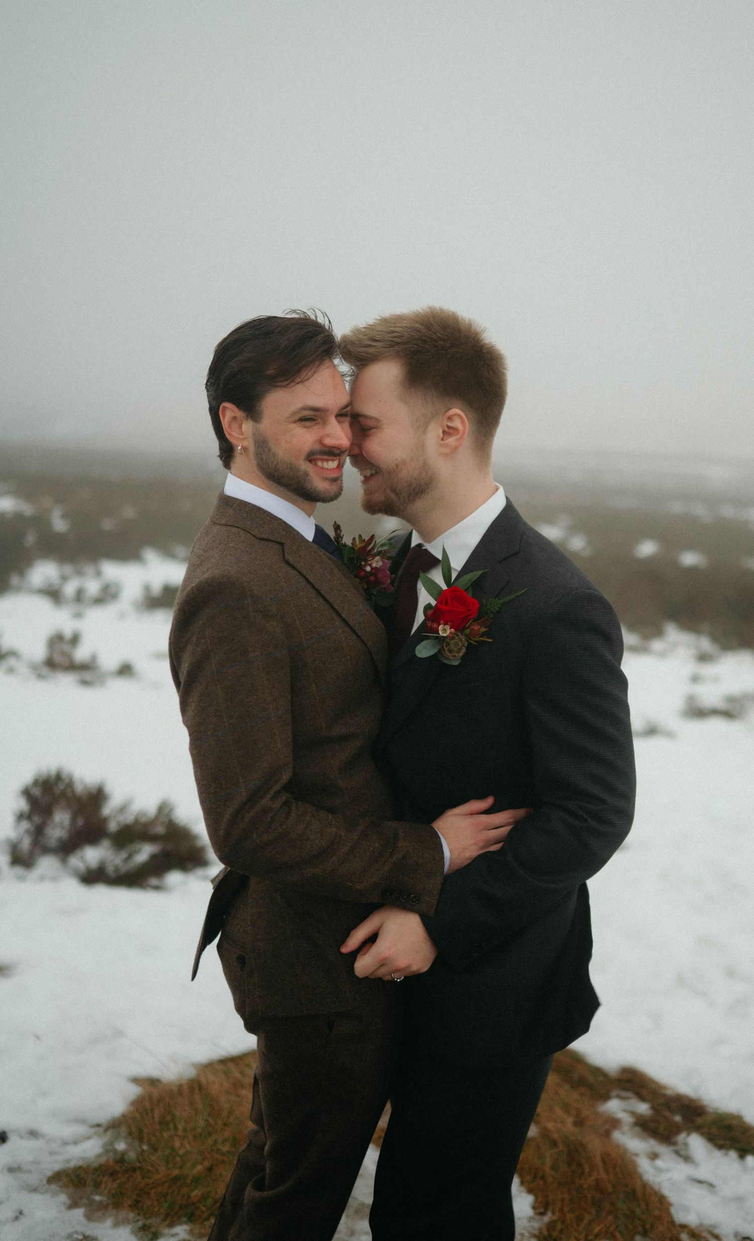 Two people in suits with red boutonnieres share an intimate moment in a snowy winter landscape.