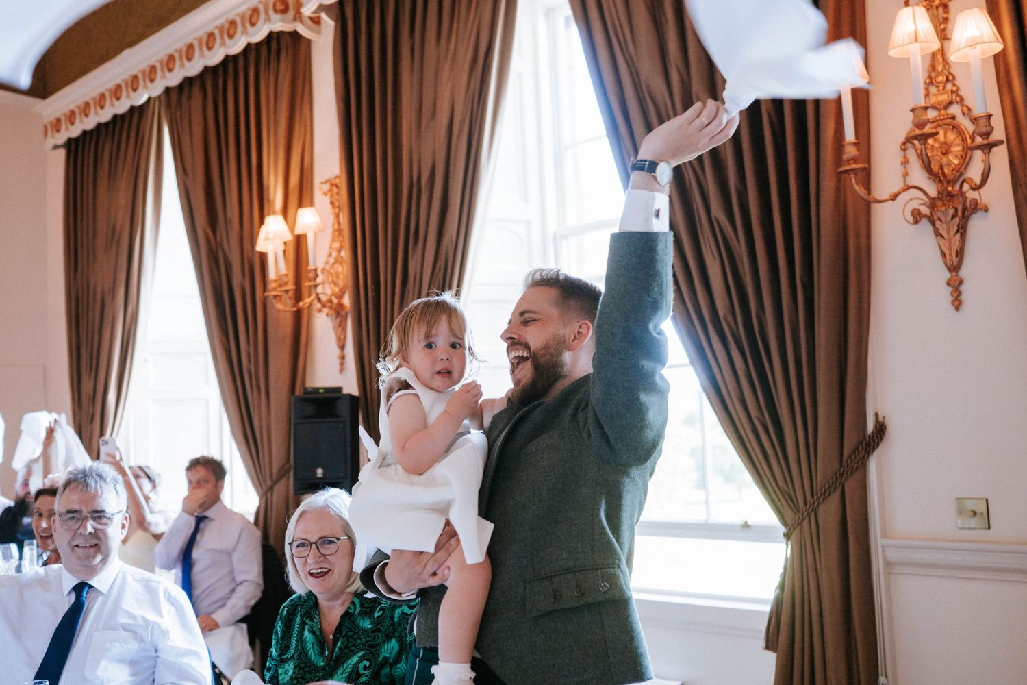 Wedding reception guests celebrating in an elegant room with brown curtains and wall sconces.
