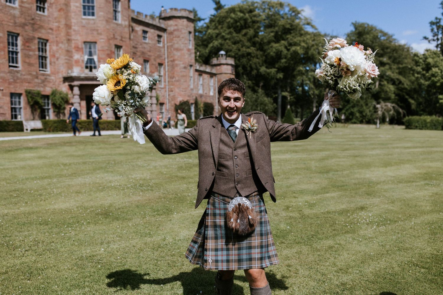 Wedding guests in traditional Scottish kilts celebrate outdoors near a historic brick building.