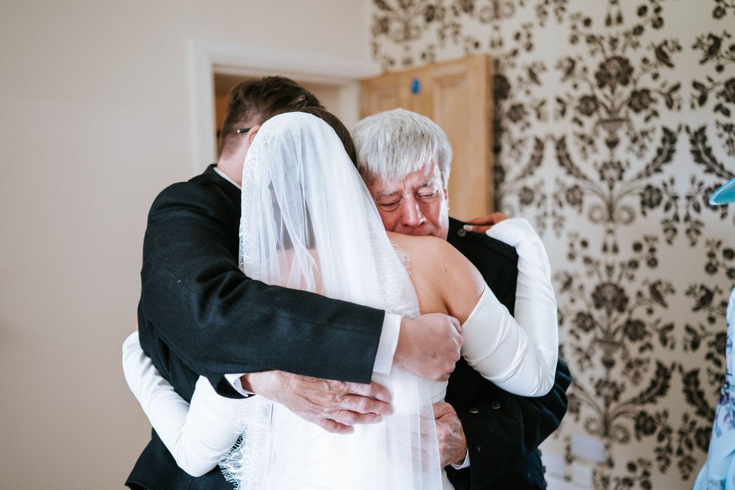 An emotional embrace between a bride in white and two others during a wedding ceremony against a floral wallpaper.