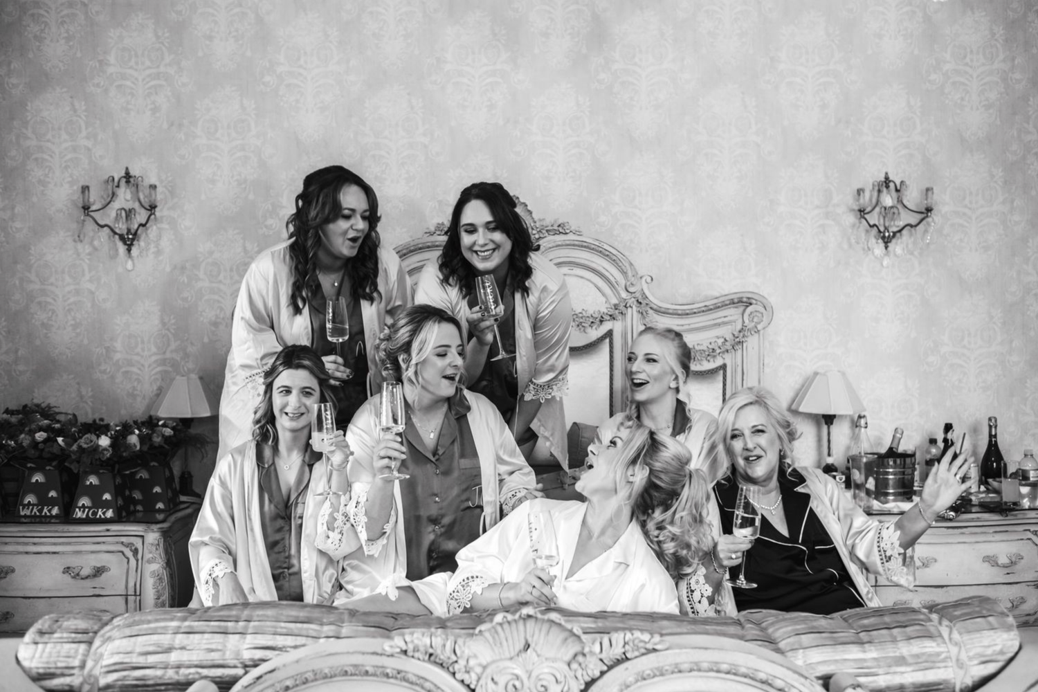 A black and white vintage style photograph shows a group lounging together on an ornate bed in matching light-colored outfits.