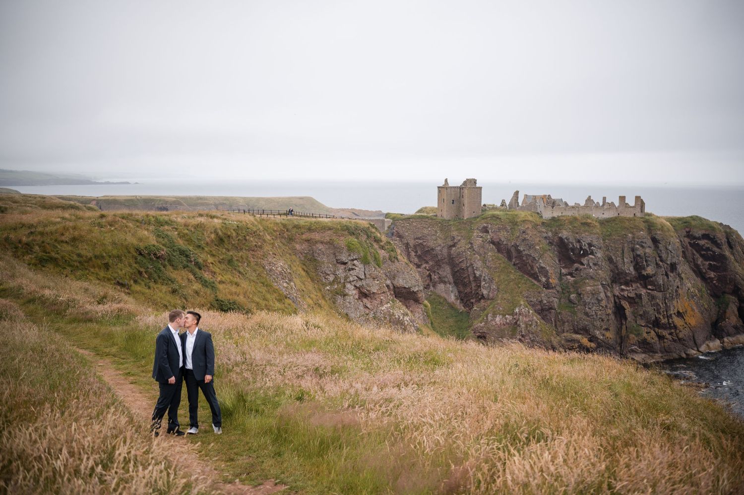 Same Sex Wedding Photography. A couple enjoys a romantic moment on a dramatic clifftop overlooking the ocean and historic castle ruins.