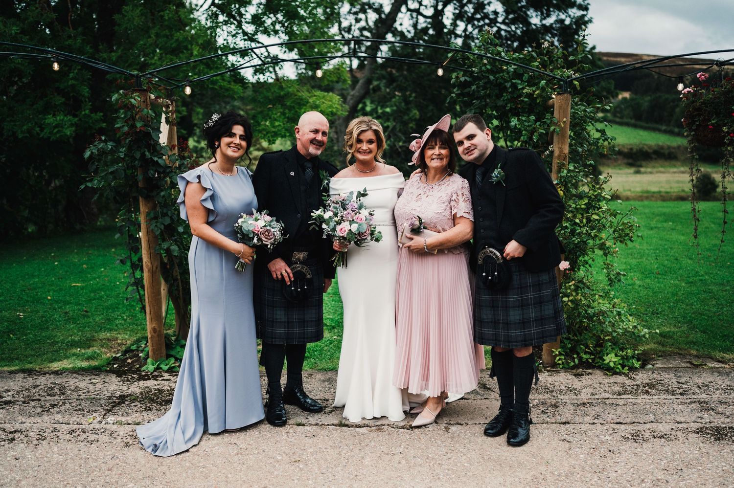 Wedding party poses outdoors in a garden setting with green trees and natural scenery.