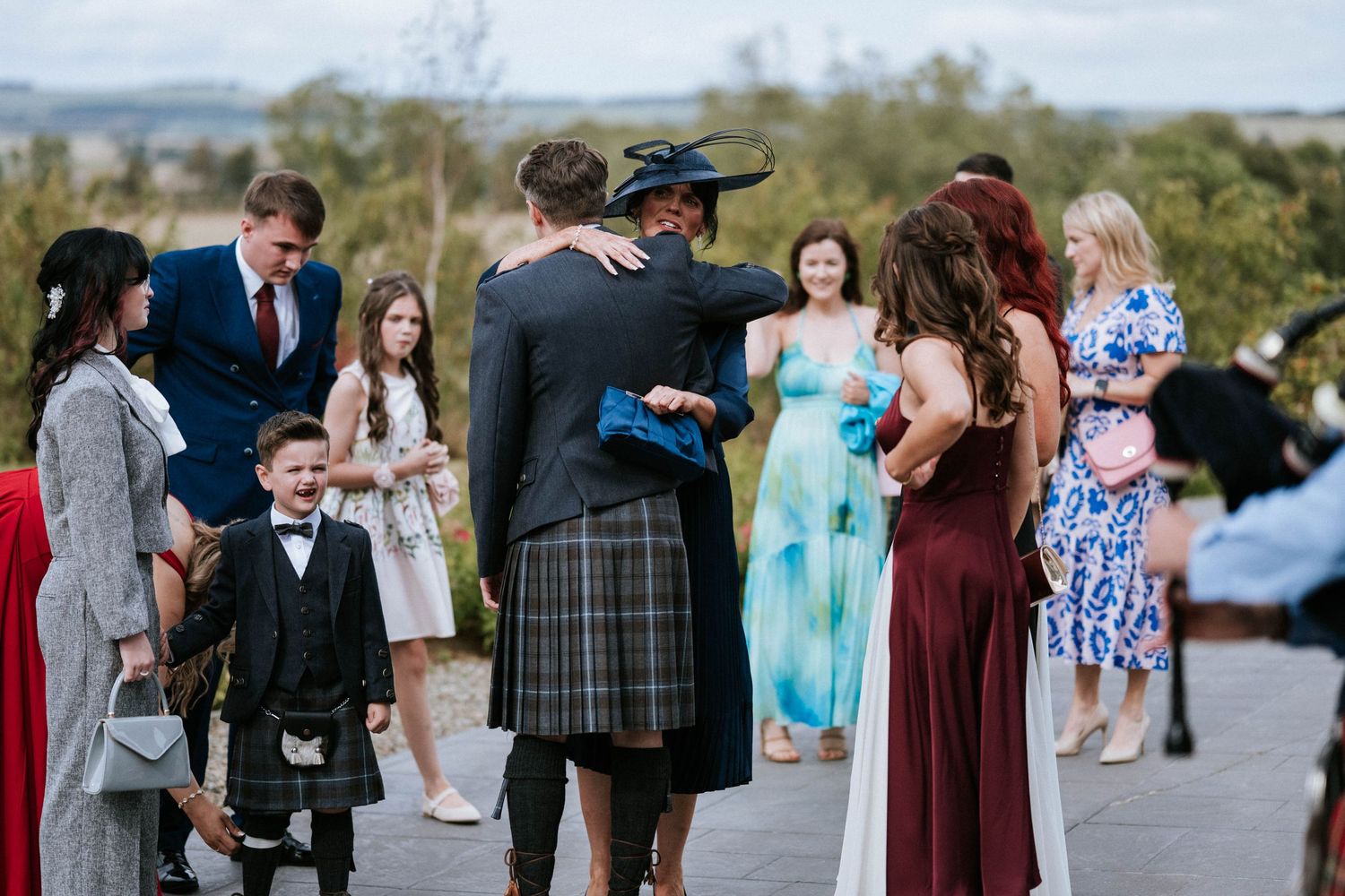 Wedding guests in kilts and formal attire gather outdoors for a Scottish celebration.