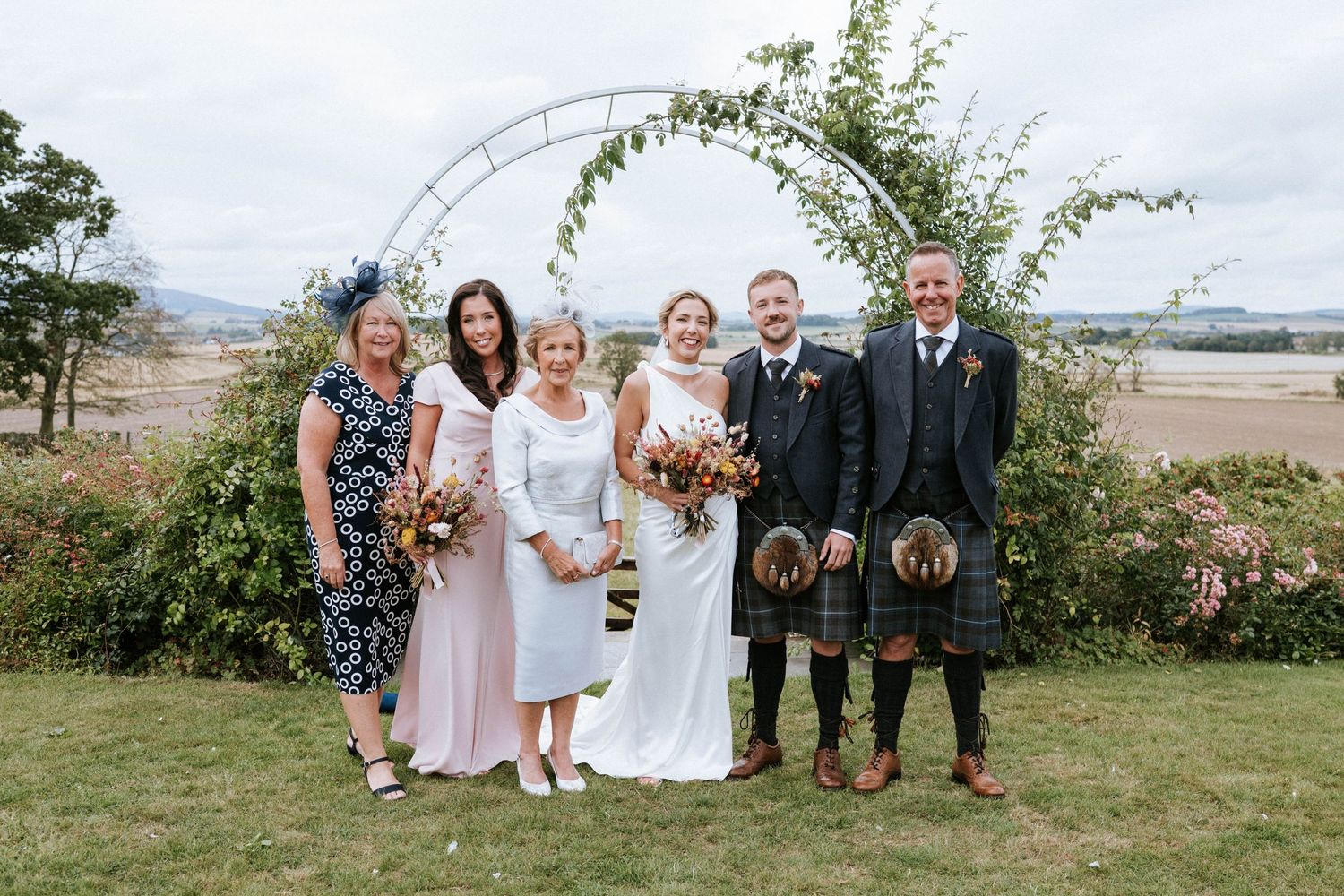 Wedding party posing for group photos in garden setting with greenery.