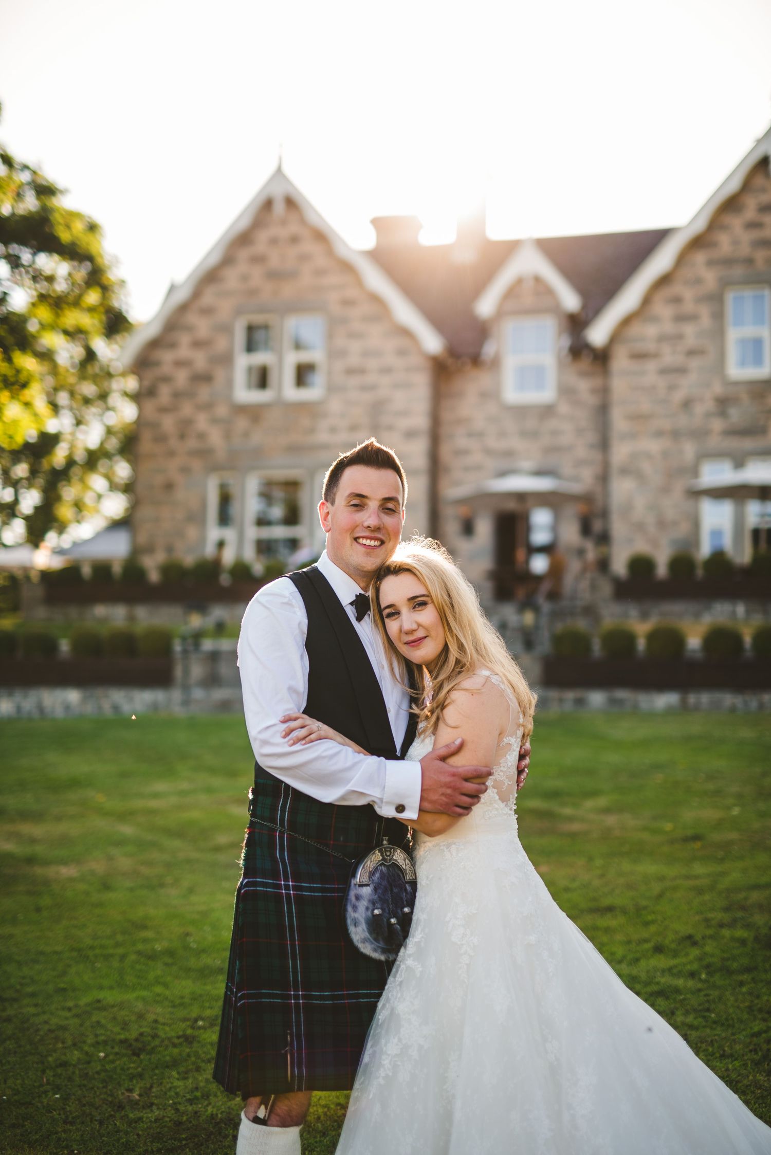 Scottish wedding couple embraces on lawn in front of stone country house at sunset.