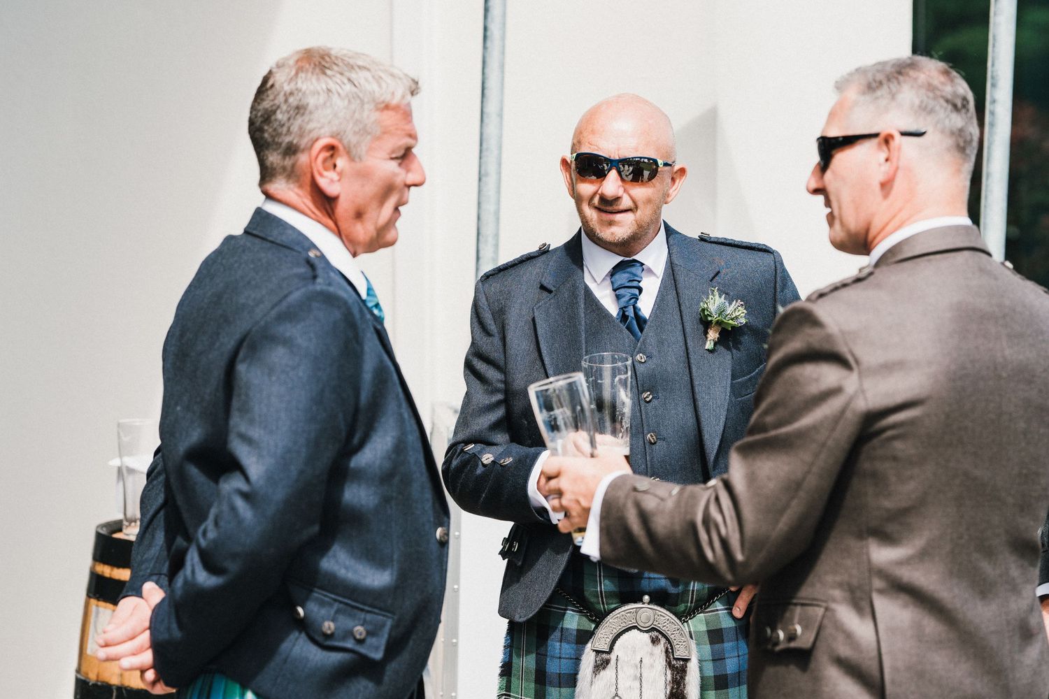 Three men in formal attire and kilts socialize with drinks at outdoor event.