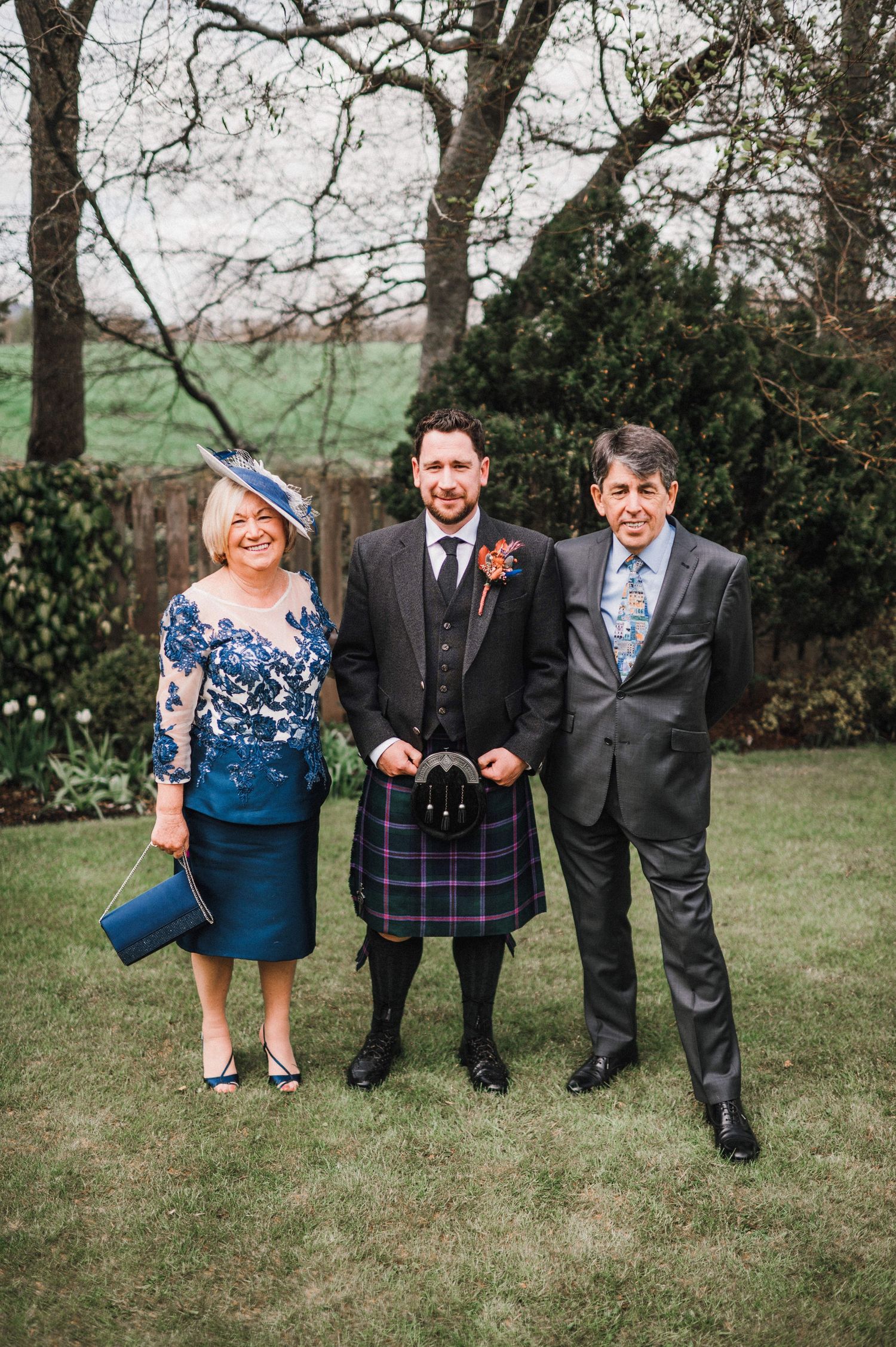 Three wedding guests pose outdoors, featuring a traditional kilt and formal attire at a Scottish celebration.