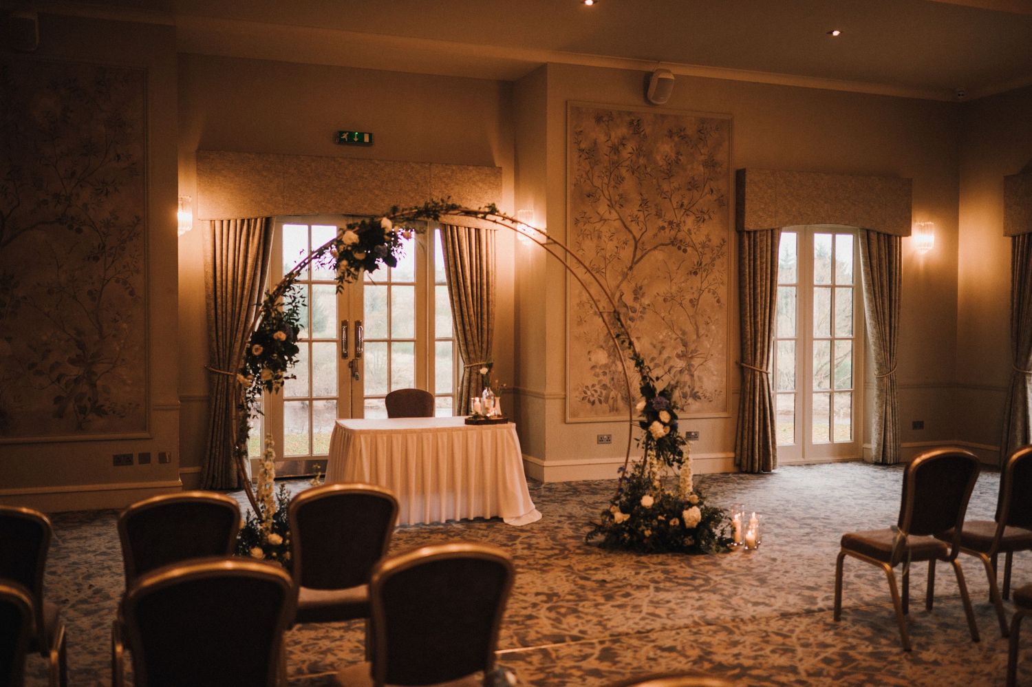 Elegant wedding venue setup with floral arch, chairs, and ornate wallpaper in warm lighting.