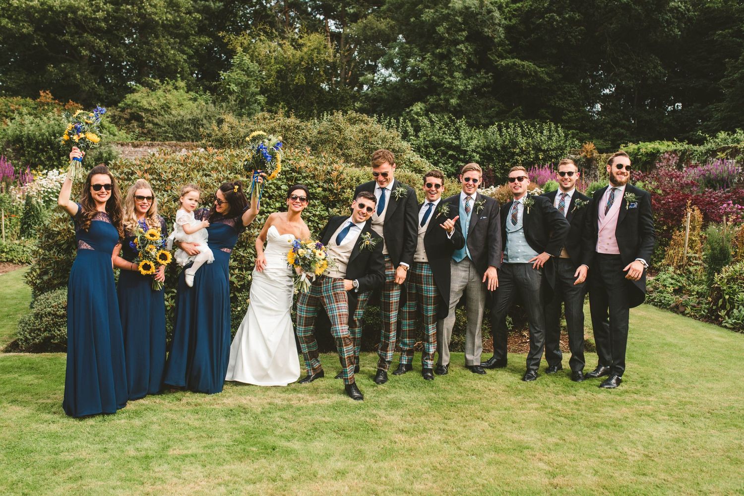 Wedding party with bridesmaids in navy and groomsmen in kilts pose on garden lawn.